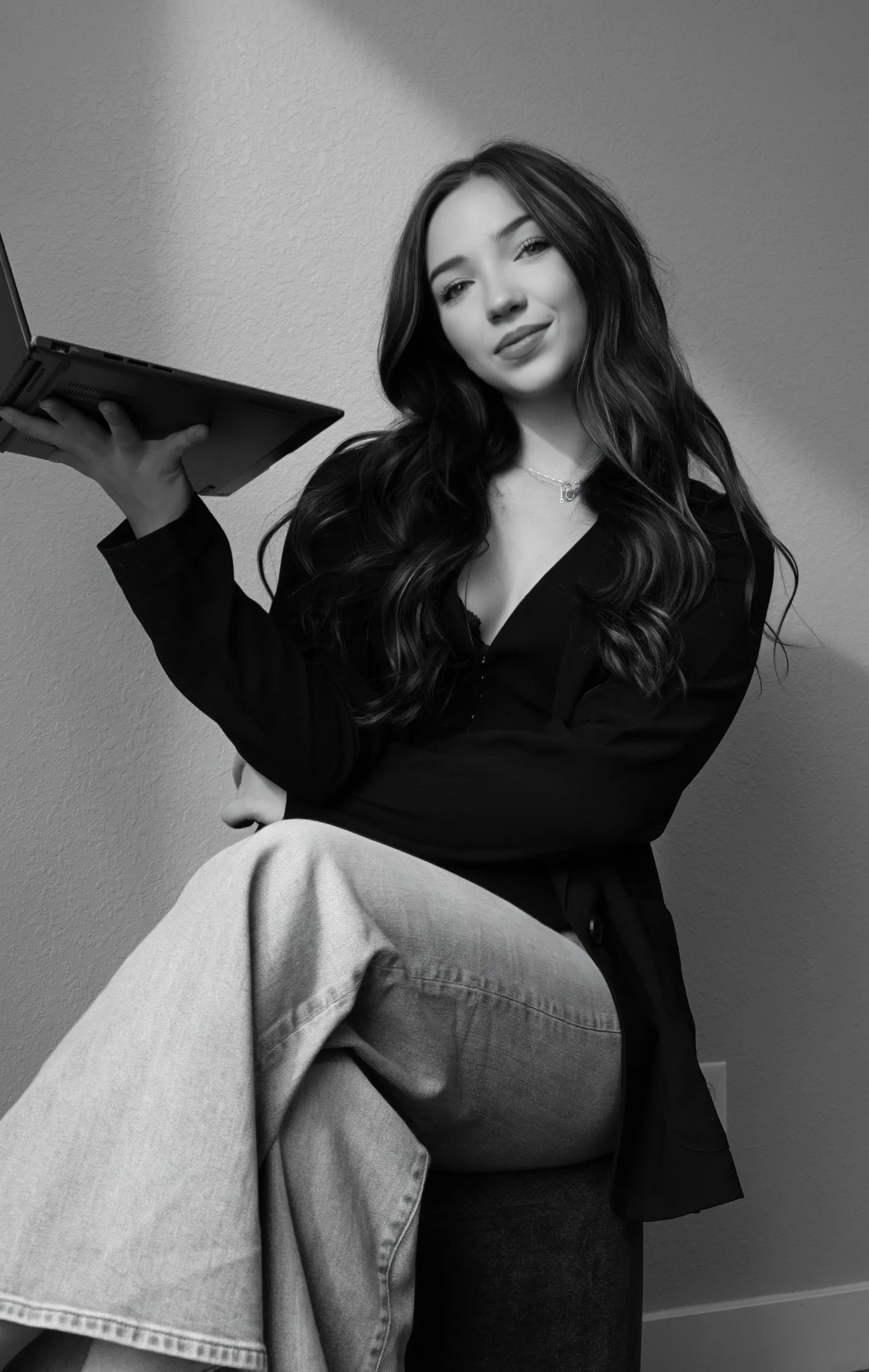 Black and white photo of a young woman with long wavy hair, sitting with one leg crossed over the other, holding a laptop in her hand. She is wearing a dark top and light-colored jeans, and is looking at the camera with a slight smile.