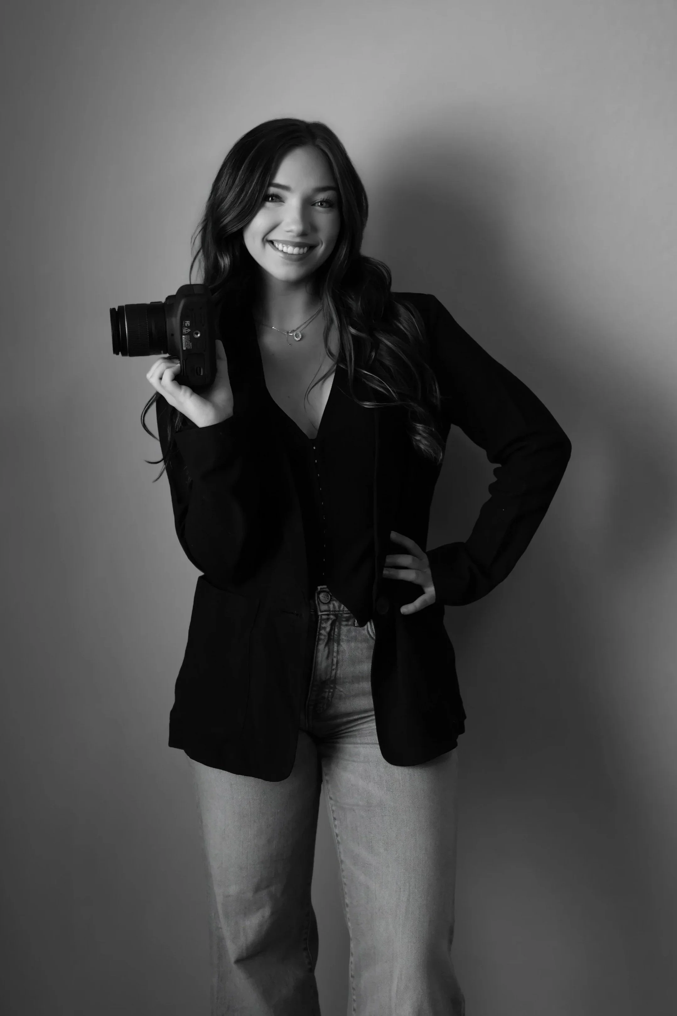 Black and white photo of a smiling woman with long hair, holding a camera on her shoulder, wearing a dark jacket and light jeans, standing against a plain backdrop.