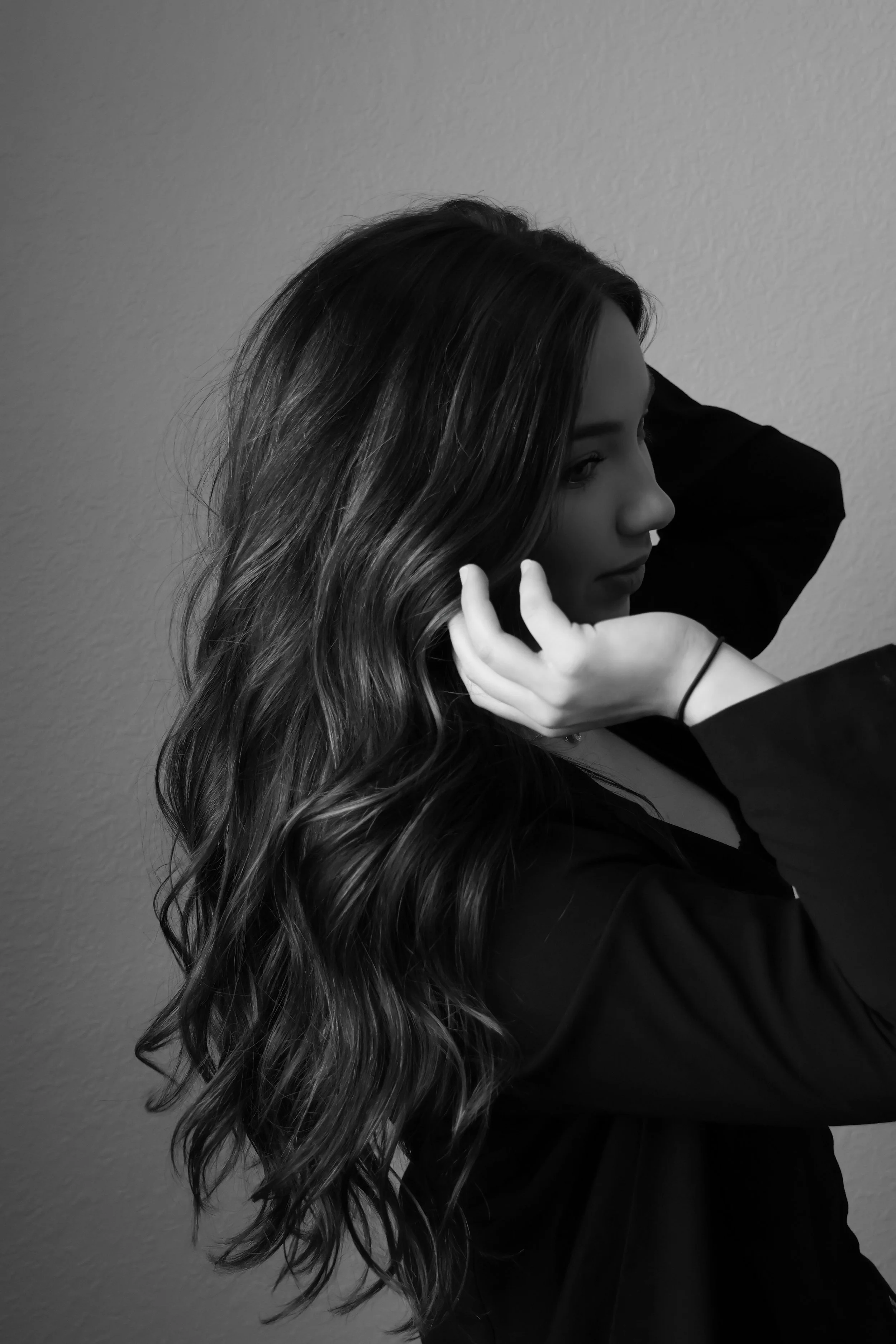 A black and white photo of a woman with long, wavy hair. She is facing right and is adjusting her hair with one hand. She is wearing a dark long-sleeved top, and her expression is contemplative. The background is a plain textured wall.