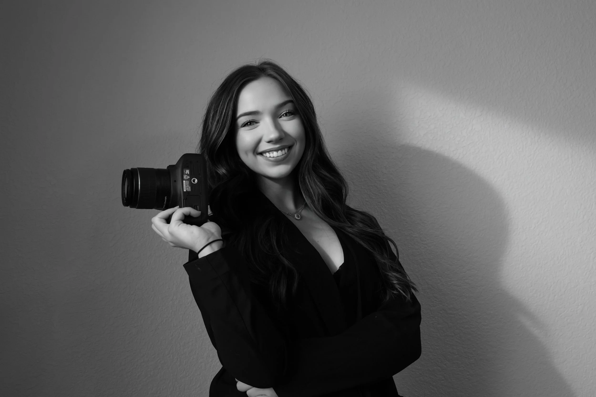 Black and white photo of a young woman with long wavy hair, smiling, holding a camera on her shoulder, standing against a plain wall with her arms crossed.
