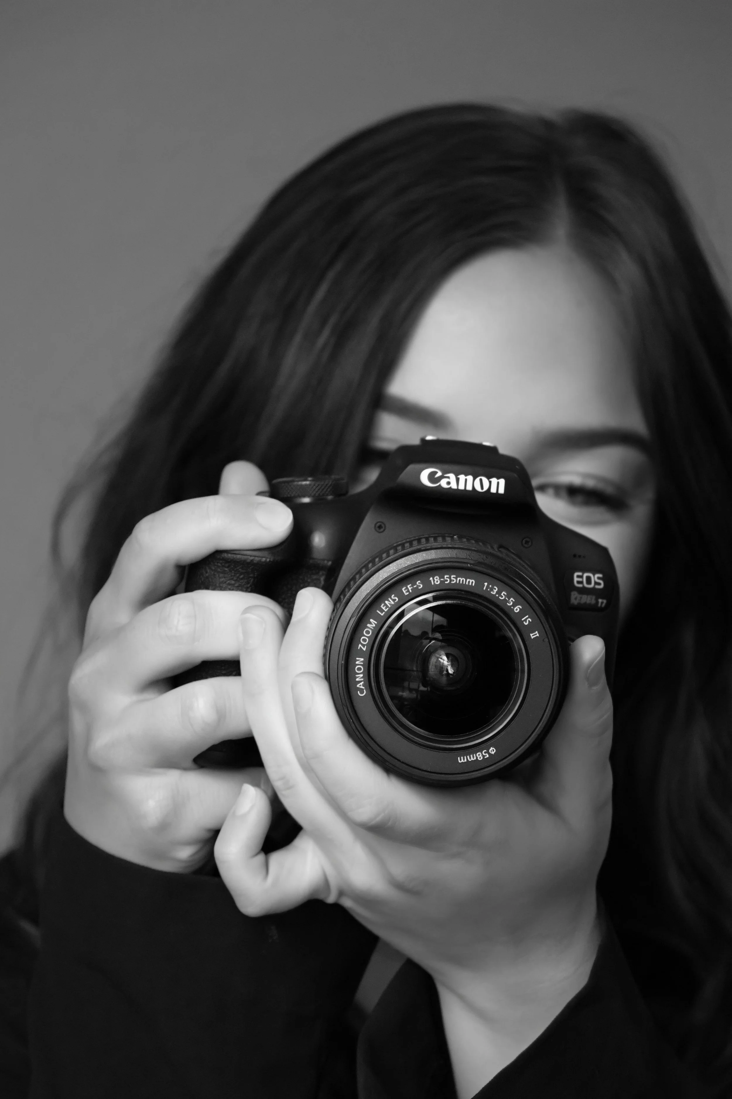A person with dark hair taking a photograph with a Canon DSLR camera, visible hand holding the camera, black and white image.