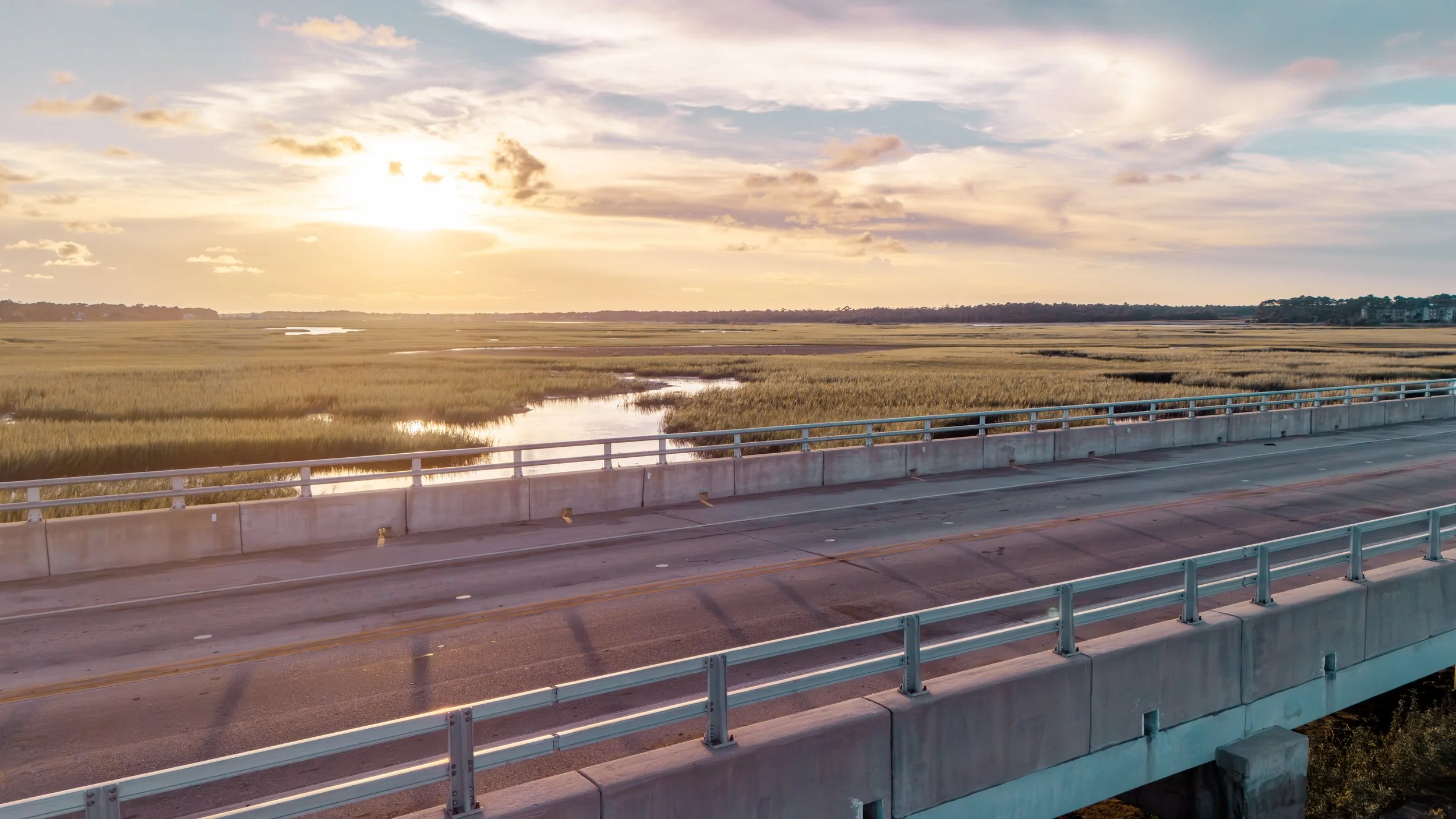 View of a bridge over marshland during sunset with partly cloudy sky.