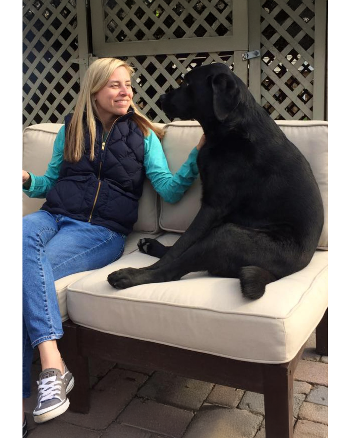 A woman sitting on a beige outdoor sofa, smiling and looking at a large black dog sitting next to her, with a lattice fence in the background.