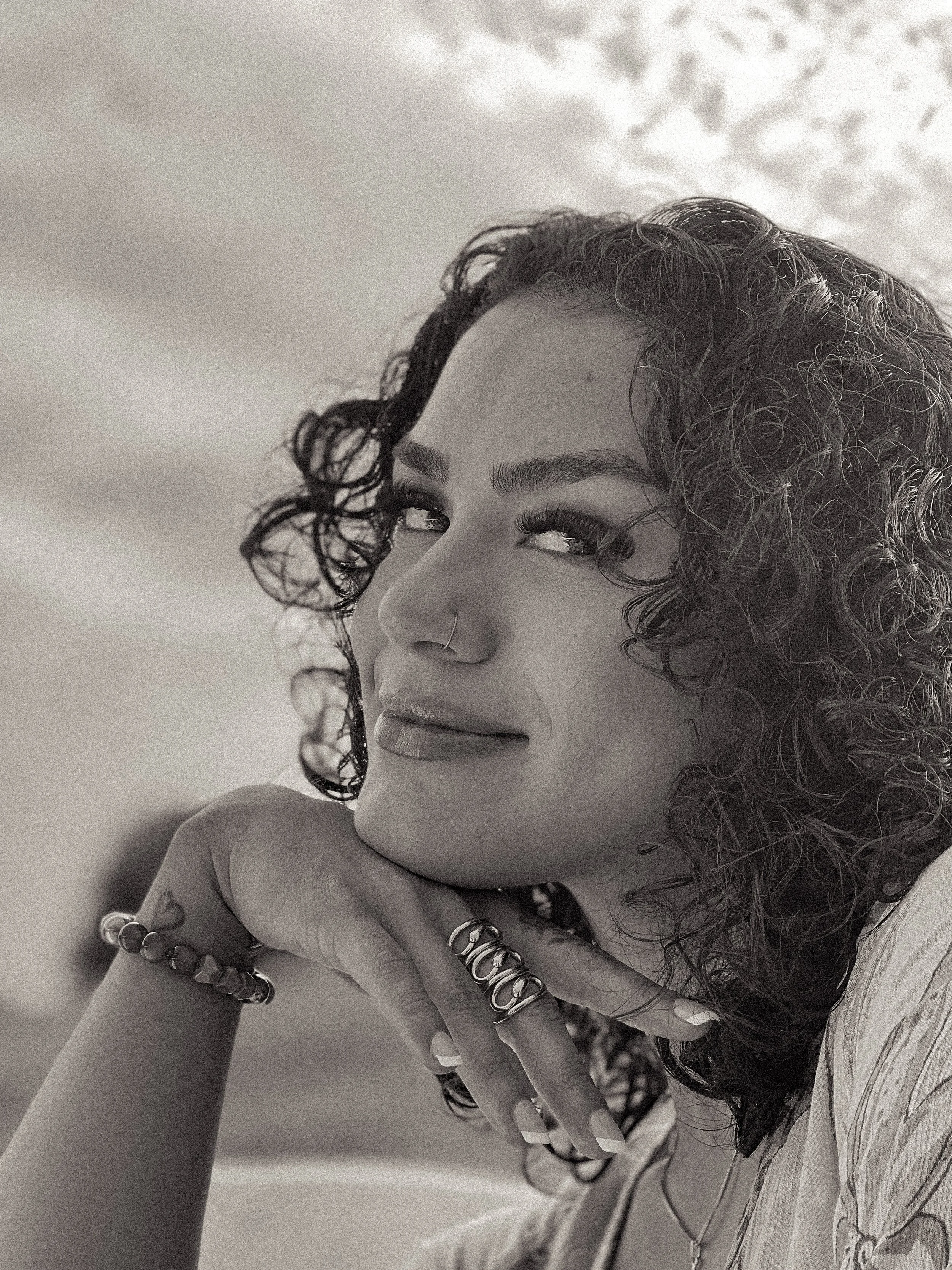 Black and white portrait of a smiling woman with curly hair, wearing rings and a nose ring, resting her chin on her hand, looking towards the camera.
