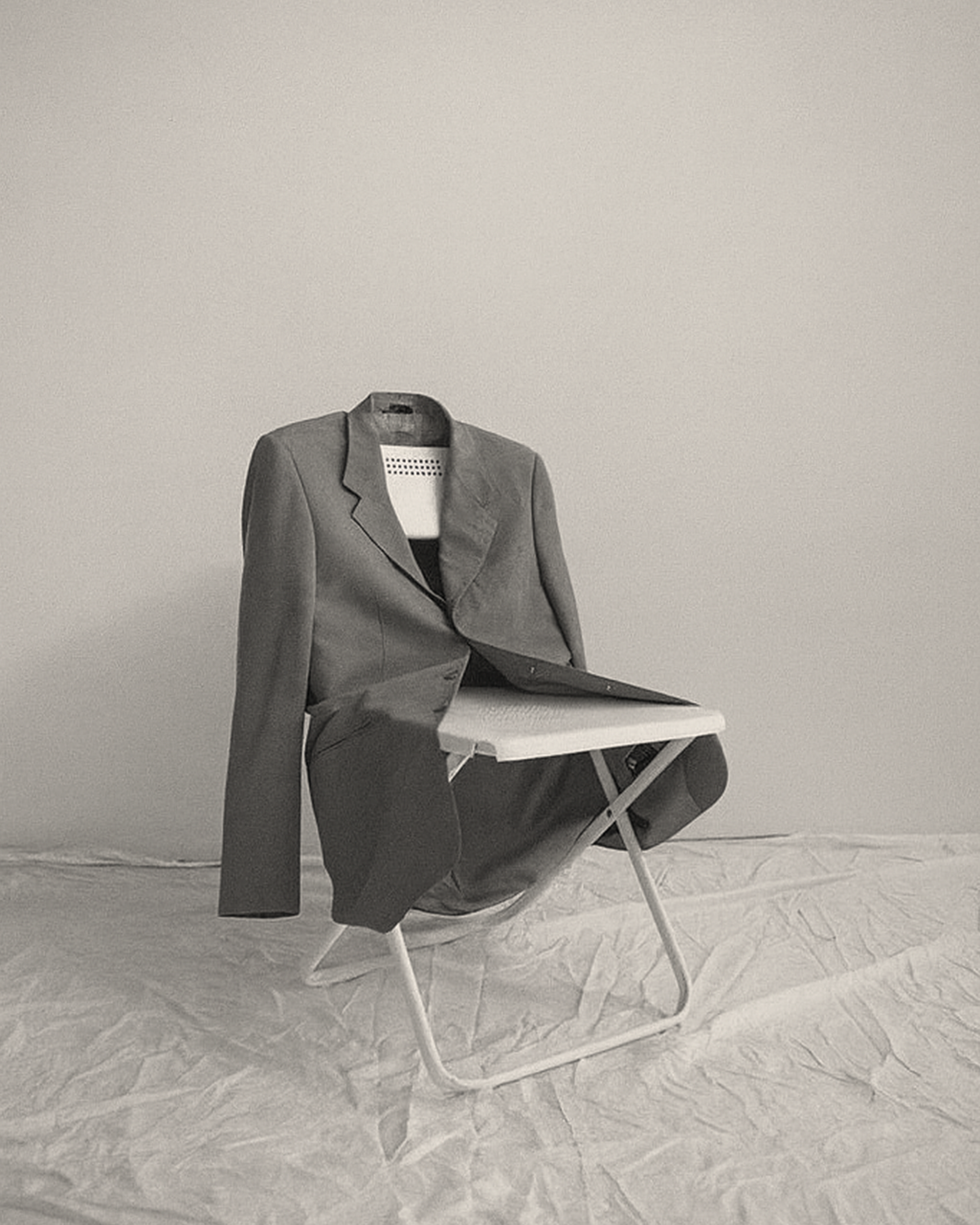 A black-and-white photo of a chair with a blazer draped over it, against a plain wall and on a crumpled cloth-covered surface.