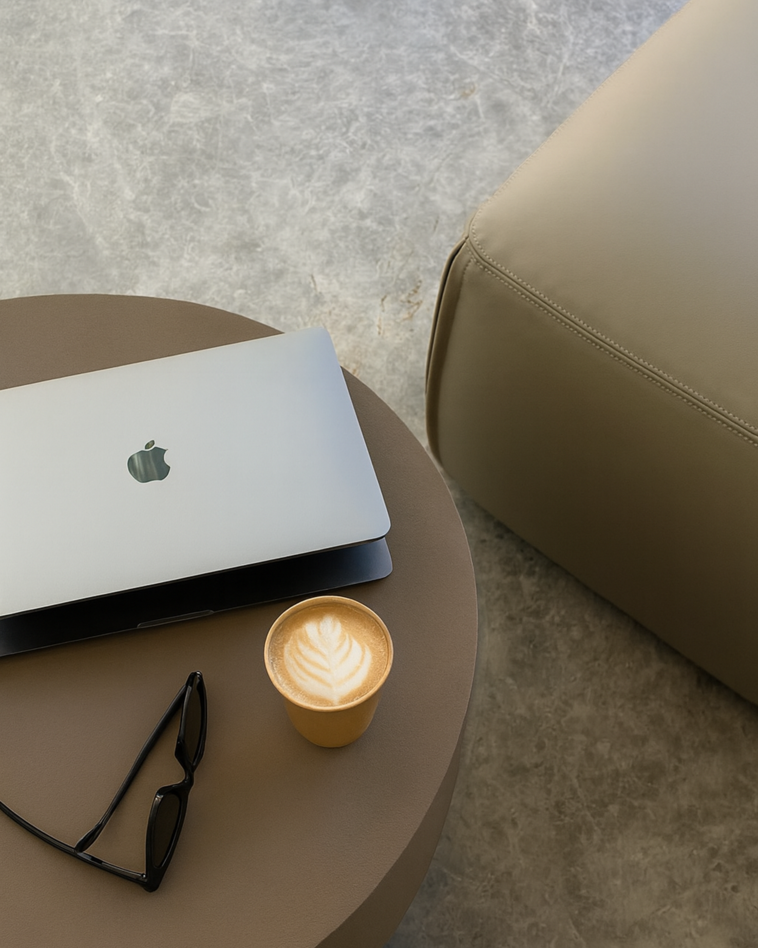 A round table with a closed laptop, a pair of black glasses, and a cup of coffee with latte art, next to a beige chair on a concrete floor.