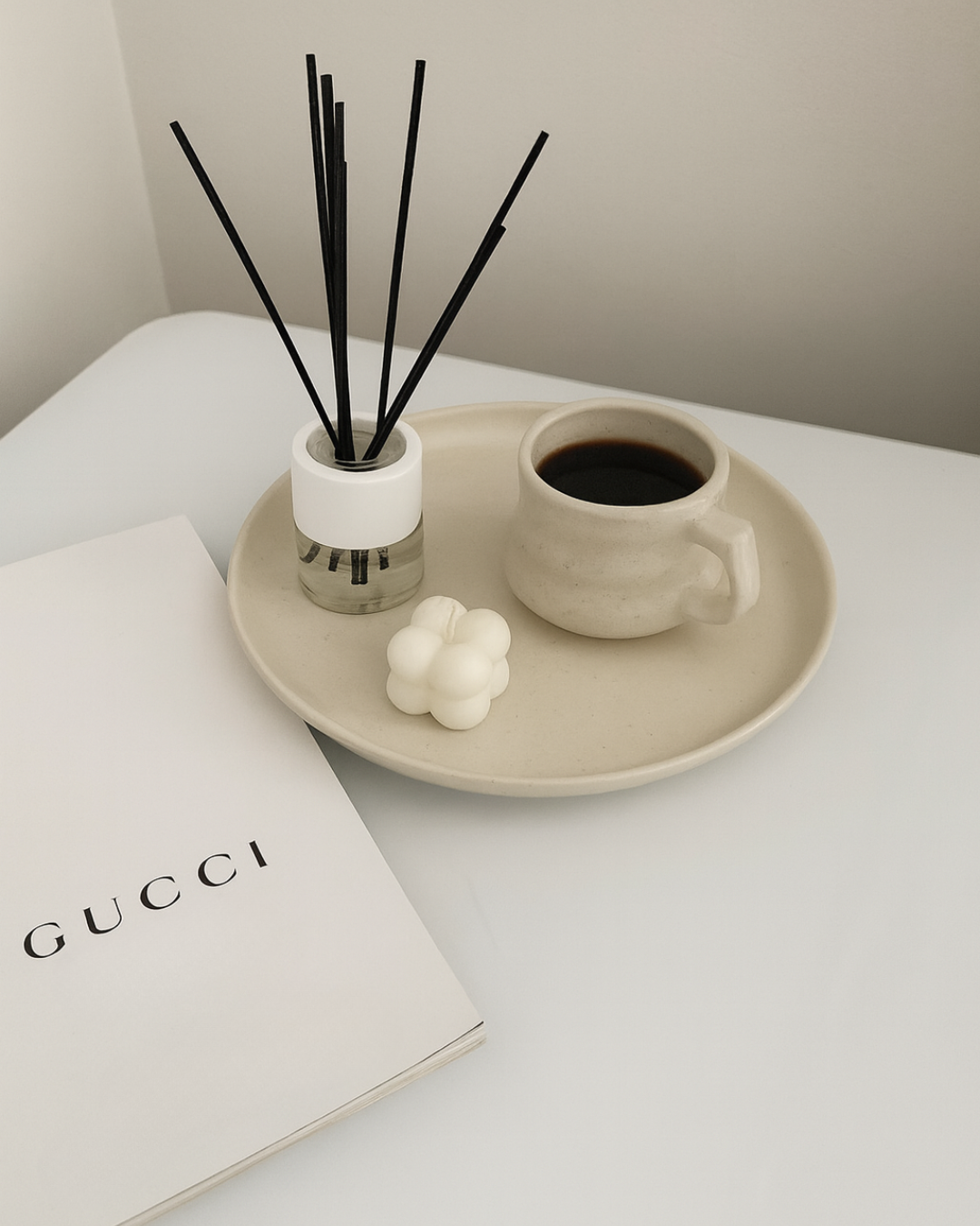 A beige ceramic tray holding a cup of black coffee, a set of black reed diffuser sticks, a white wax candle in the shape of a cluster of bubbles, and a small white booklet titled 'GUCCI' on a white surface.