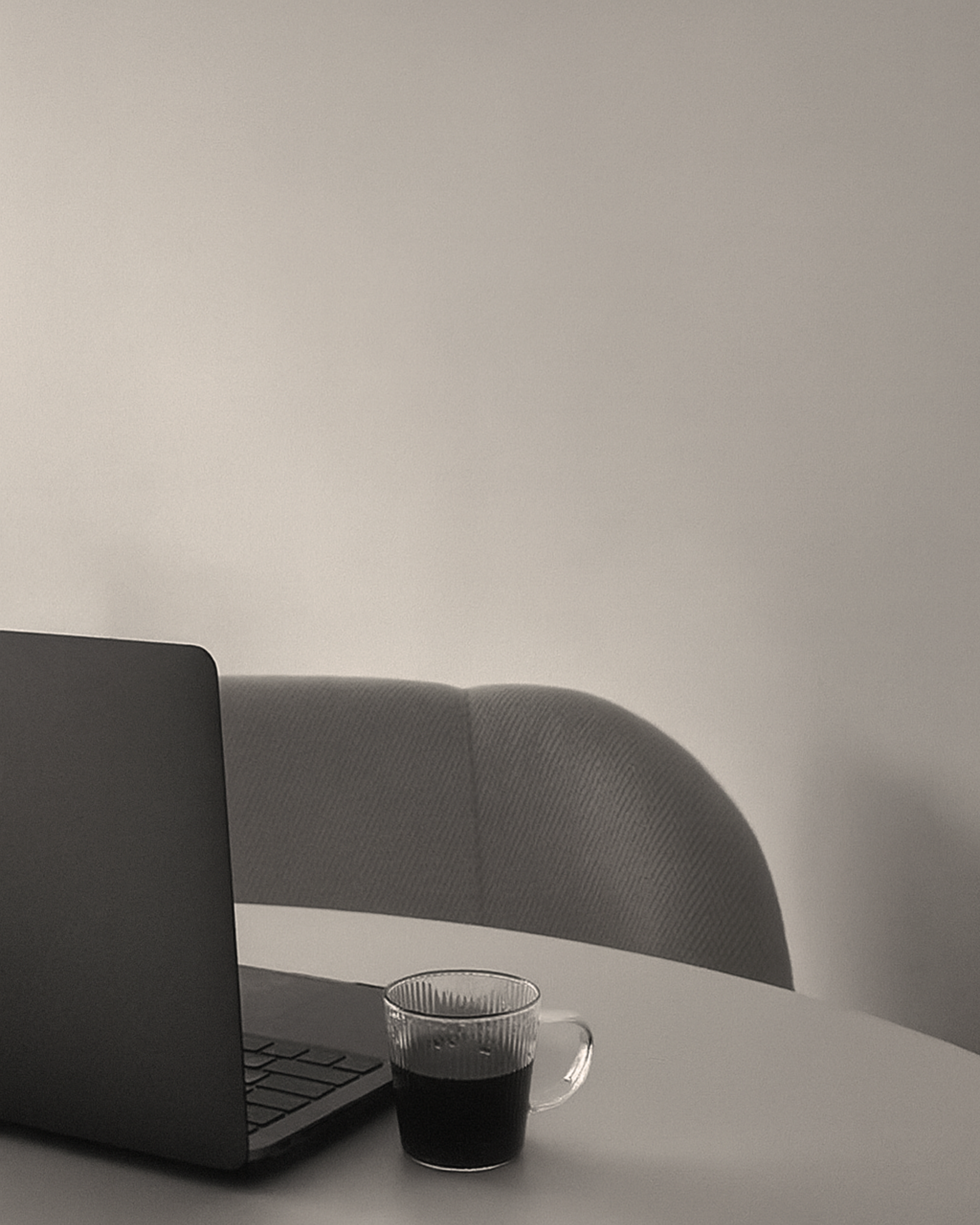 Laptop on a white table with a glass mug of dark coffee or tea, with a textured grey chair in the background.
