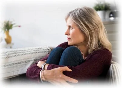 A woman sitting on a couch with her knees drawn up and arms wrapped around her legs, looking troubled with no obvious solutions.