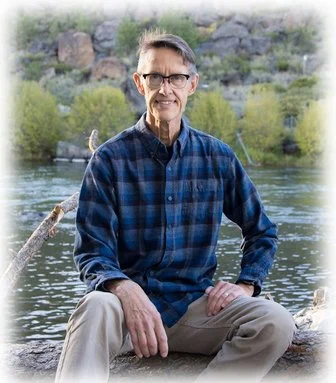 A supportive looking man with glasses, wearing a blue plaid shirt, sitting on a rock by a river with trees and rocks in the background.