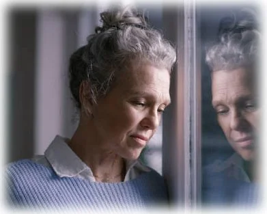 An elderly woman sees her reflection in a window.  She appears despondent, obviously in grief.
