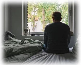 Despondent person sitting on bed and looking hopelessly out of a window.
