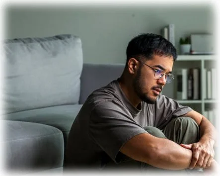 A man with his arms crossed sitting on the floor of his living room, looking down with a serious expression. He appears conflicted.