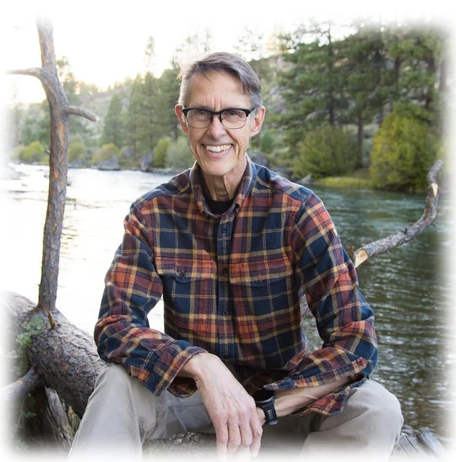 A smiling man in glasses and a plaid shirt sitting outdoors near a river with trees in the background.