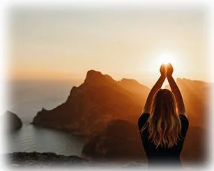 A woman standing on a rocky coastline during sunset, holding her hands together above her head as if to hold the sun.