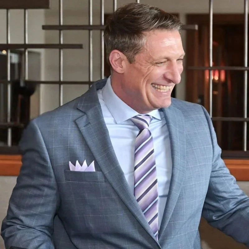 A man in a gray checkered suit, white dress shirt, and striped tie, smiling and looking to his right, in an indoor setting with wooden and metal decor.