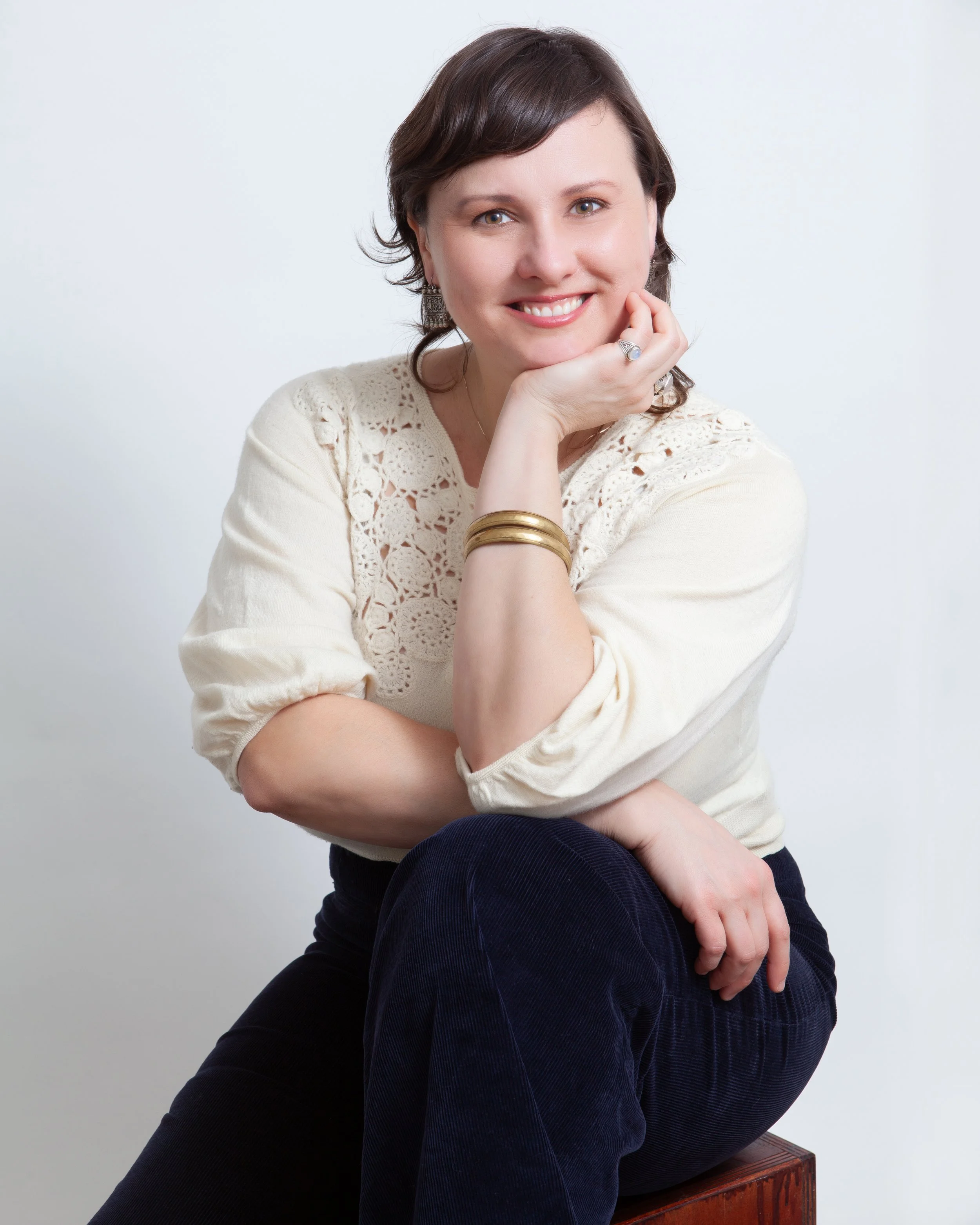 Portrait of a smiling woman with short dark hair, wearing a cream-colored crochet top and dark pants, sitting with her elbow on her knee, against a plain white background.