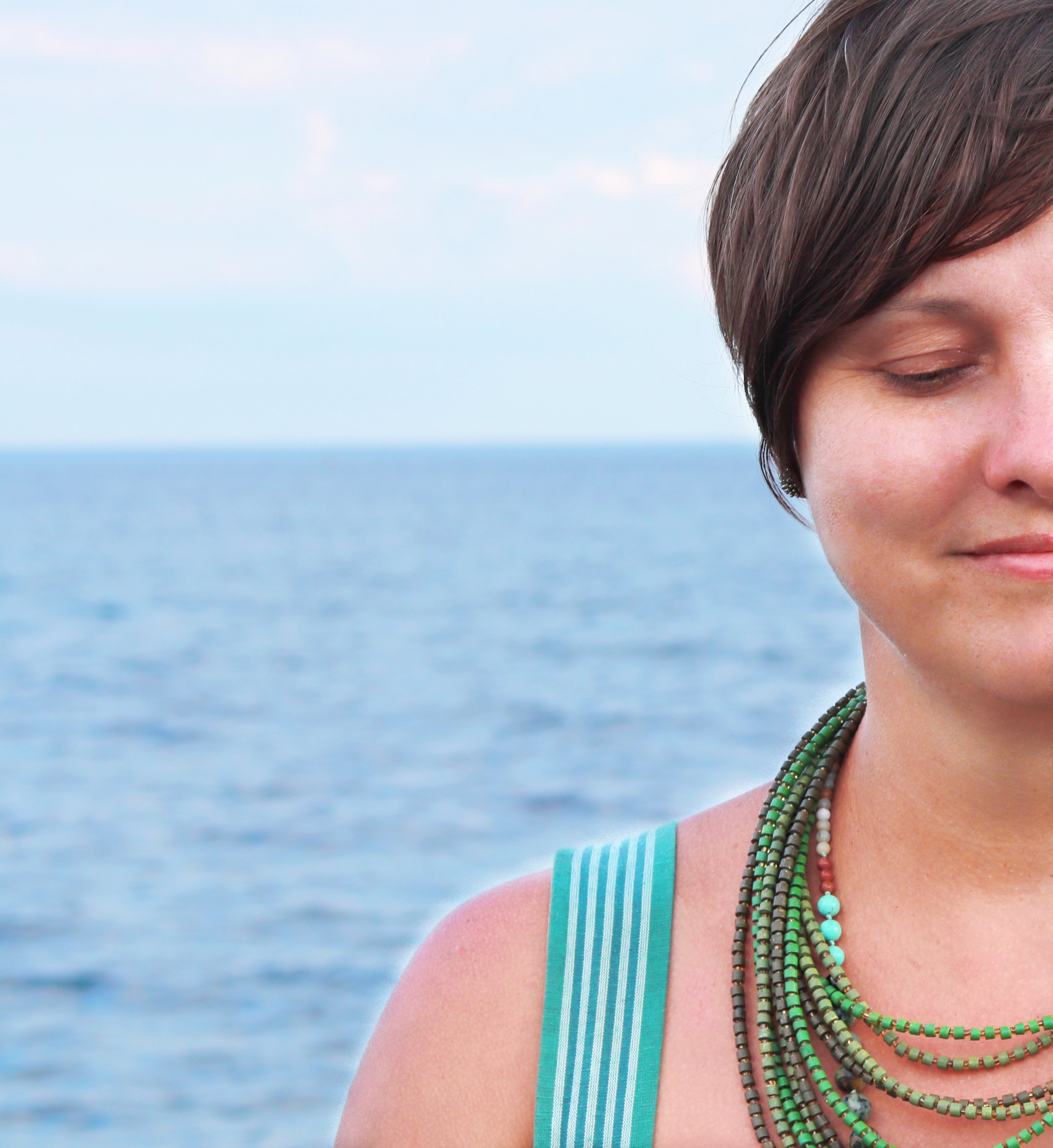 Close-up of a woman with short brown hair, wearing multiple beaded necklaces, smiling softly with her eyes closed, against a background of the ocean and blue sky.