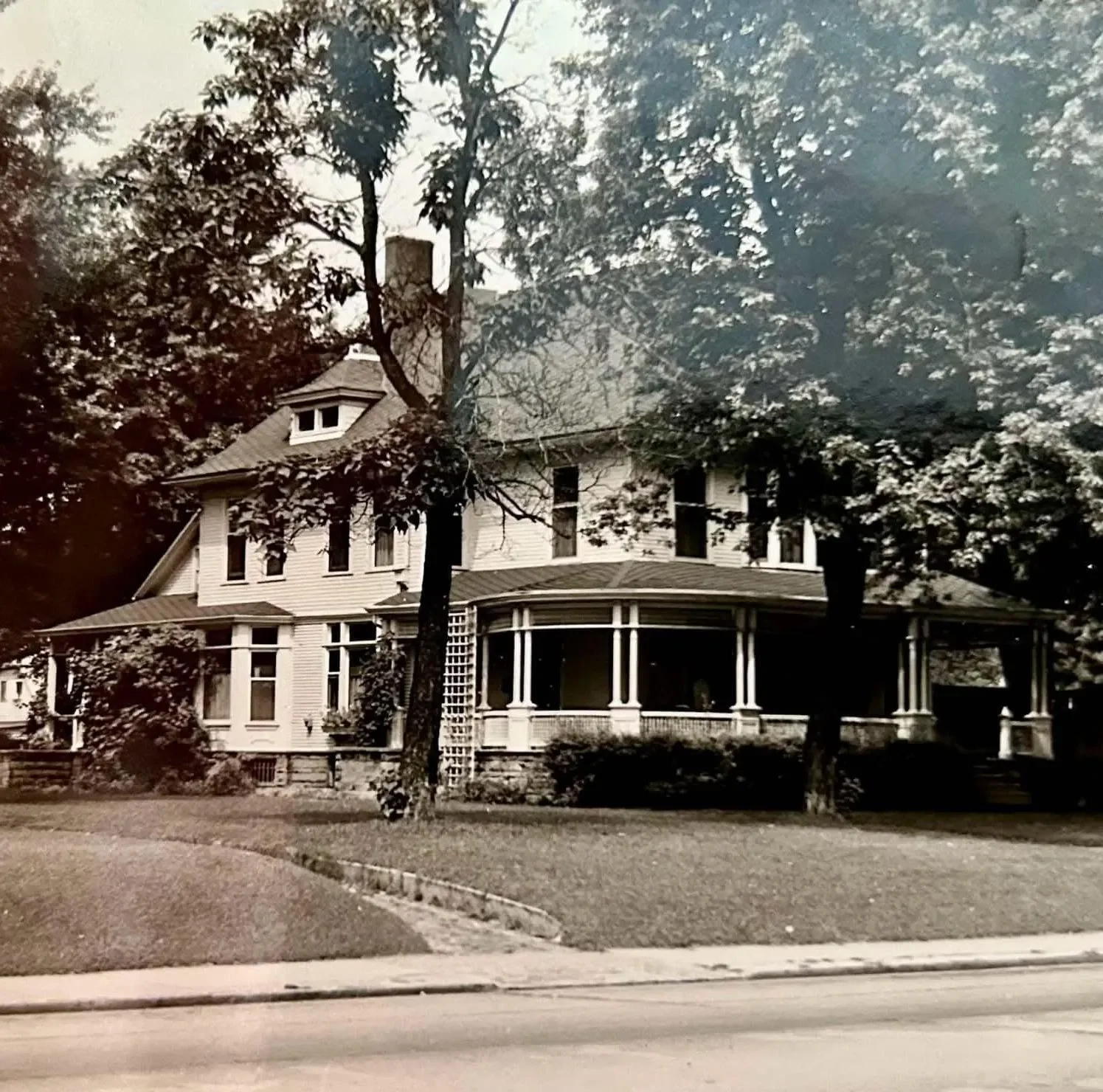 A large, old-fashioned, white Victorian house with a wraparound porch and multiple stories, surrounded by trees on a residential street.