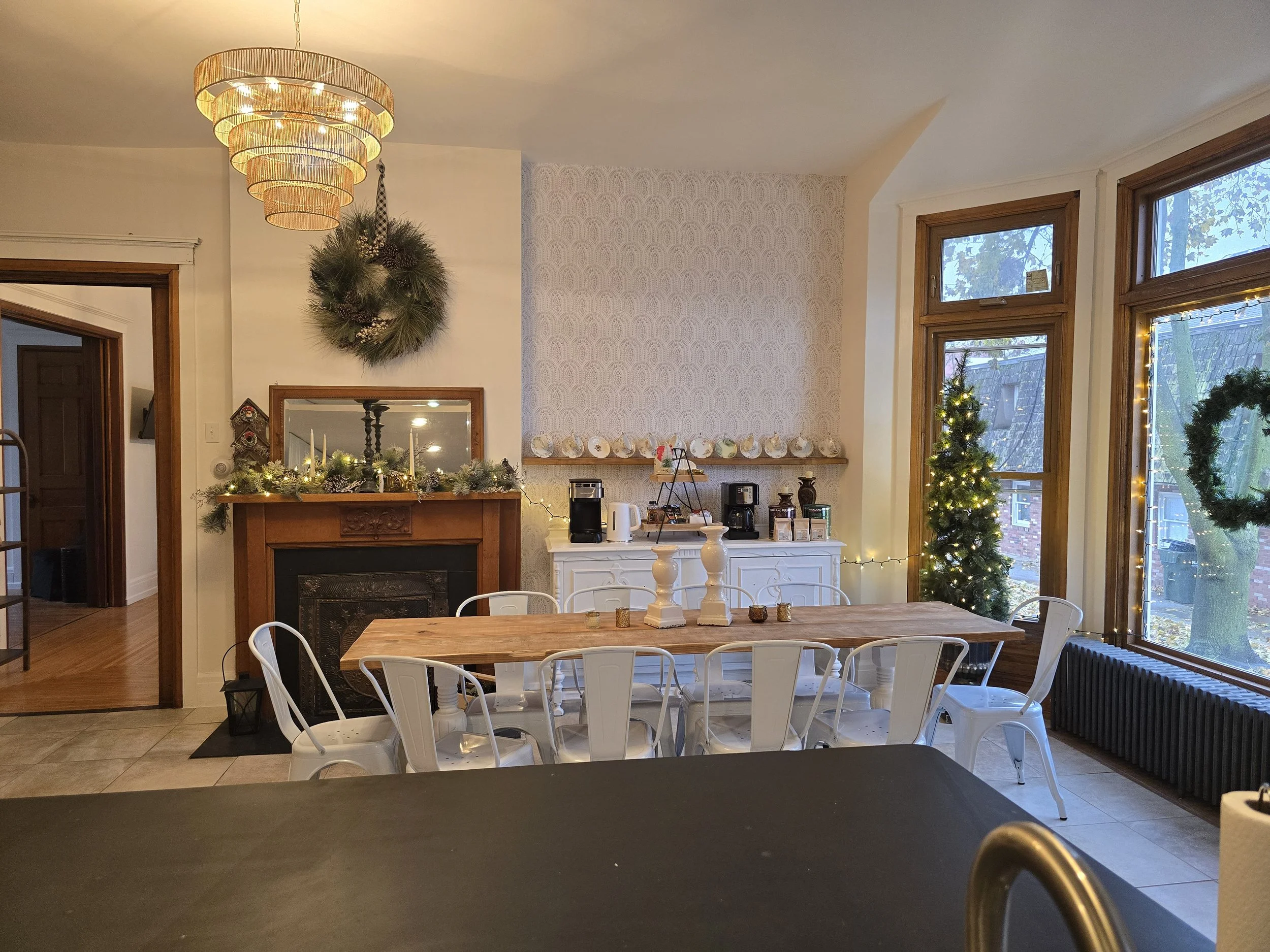 A dining area decorated for Christmas featuring a wooden table with white chairs, a small decorated Christmas tree, a fireplace with greenery and candles, a wreath on the wall, large windows with fairy lights, and a sideboard with coffee-making appliances.