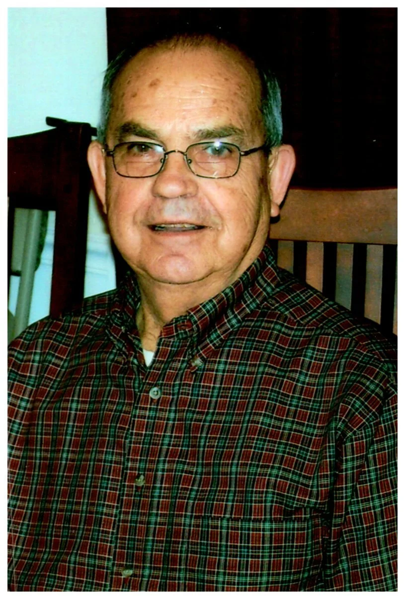 An elderly man with glasses wearing a red and green plaid shirt, sitting in a wooden chair, smiling at the camera.