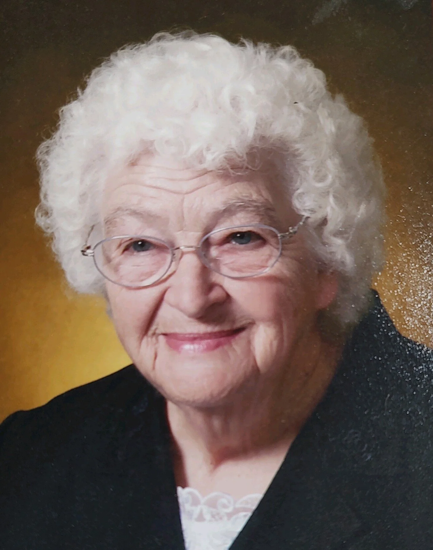 An elderly woman with curly white hair, wearing glasses, smiling, dressed in a black suit with a white lace top, sitting against a gold-colored background.