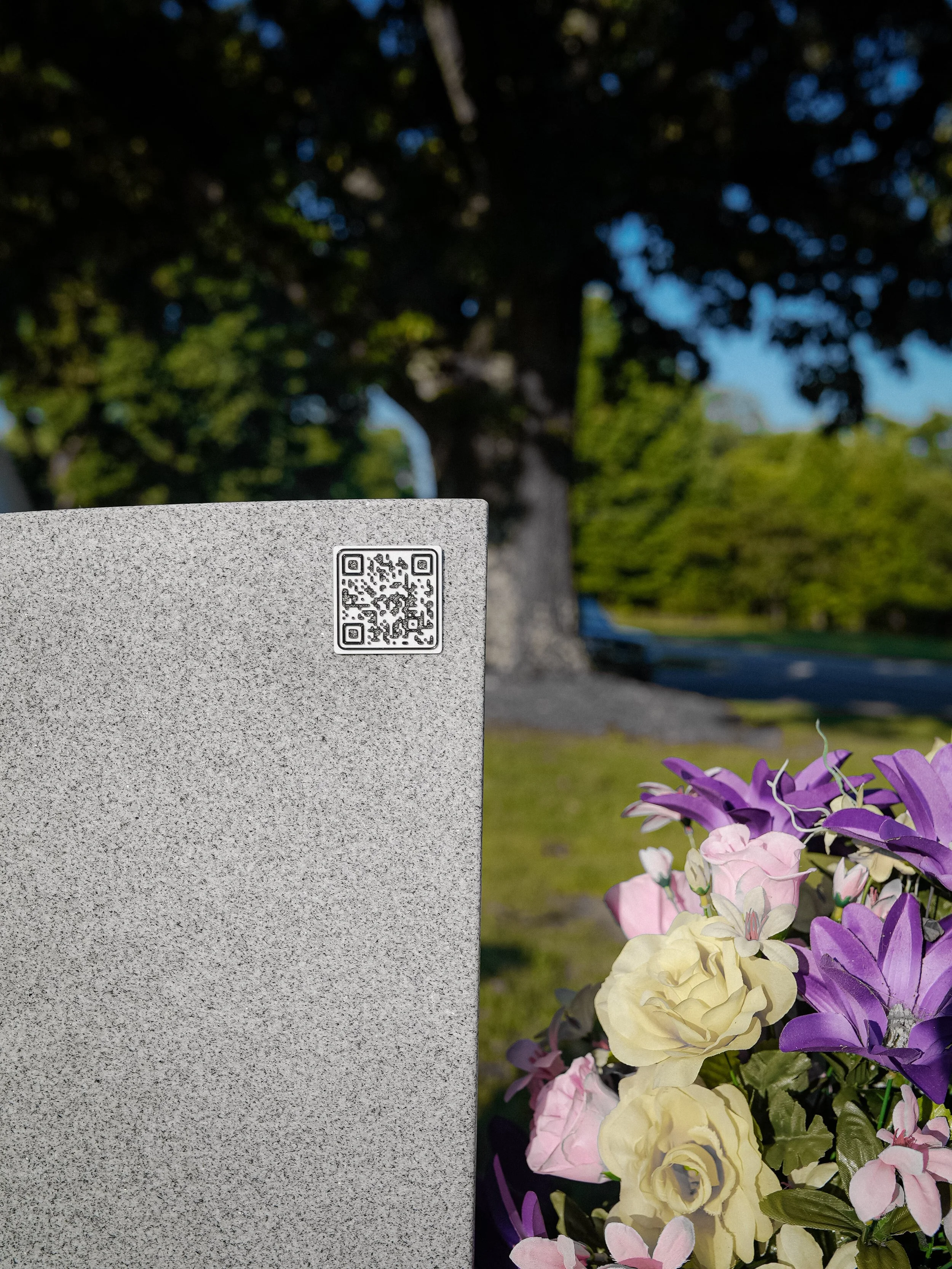 A flower arrangement with pink, purple, and cream artificial flowers next to a gray marker with a QR code, set outdoors with trees and a blue sky in the background.