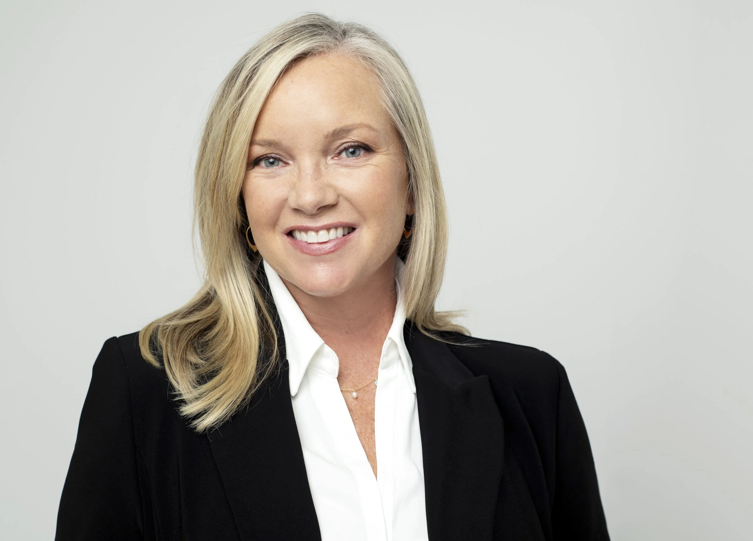 A smiling woman with blonde hair wearing a black blazer and white shirt against a plain background.