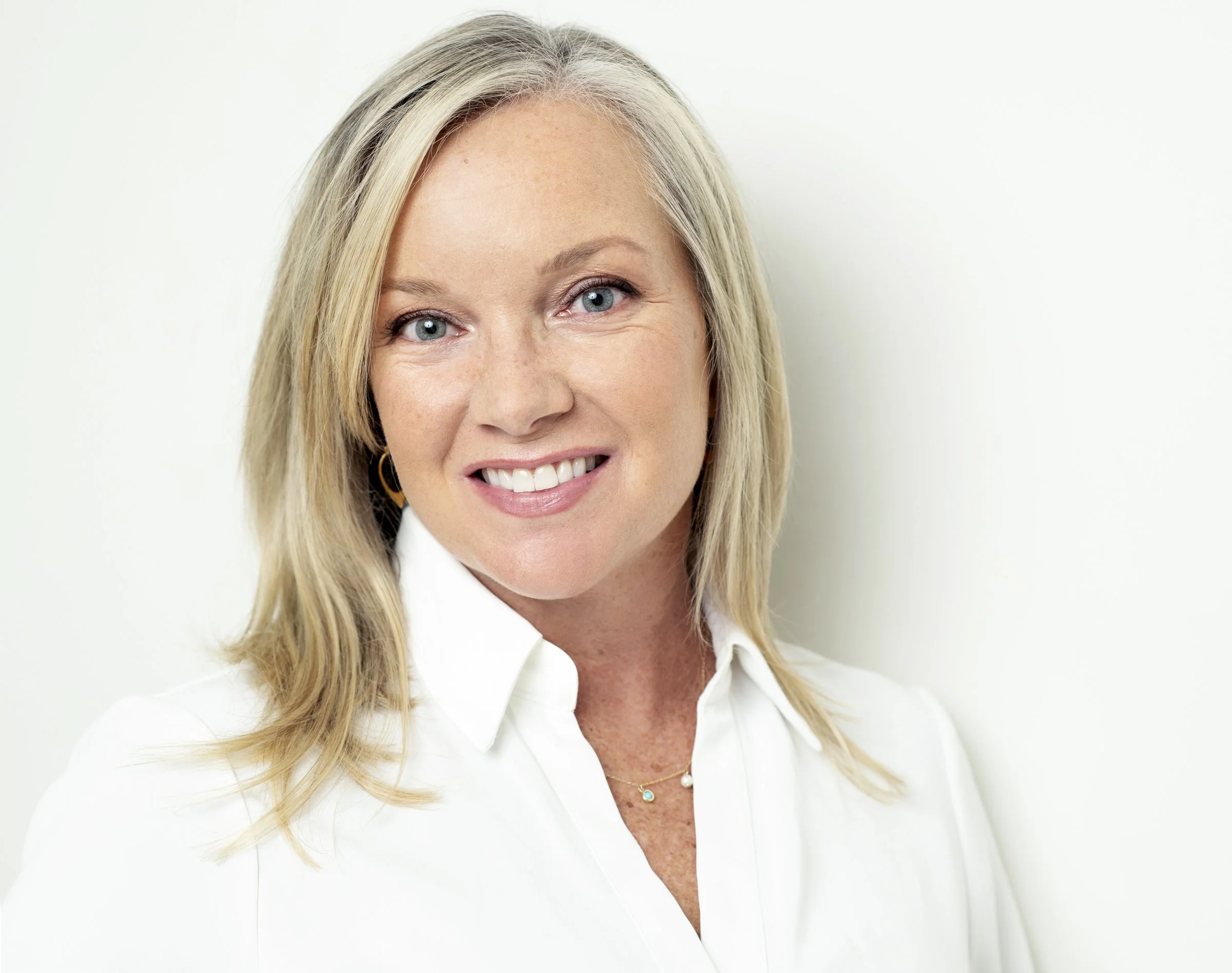 Head and shoulders portrait of a smiling woman with blonde hair and blue eyes, wearing a white collared shirt and gold jewelry, against a white background.