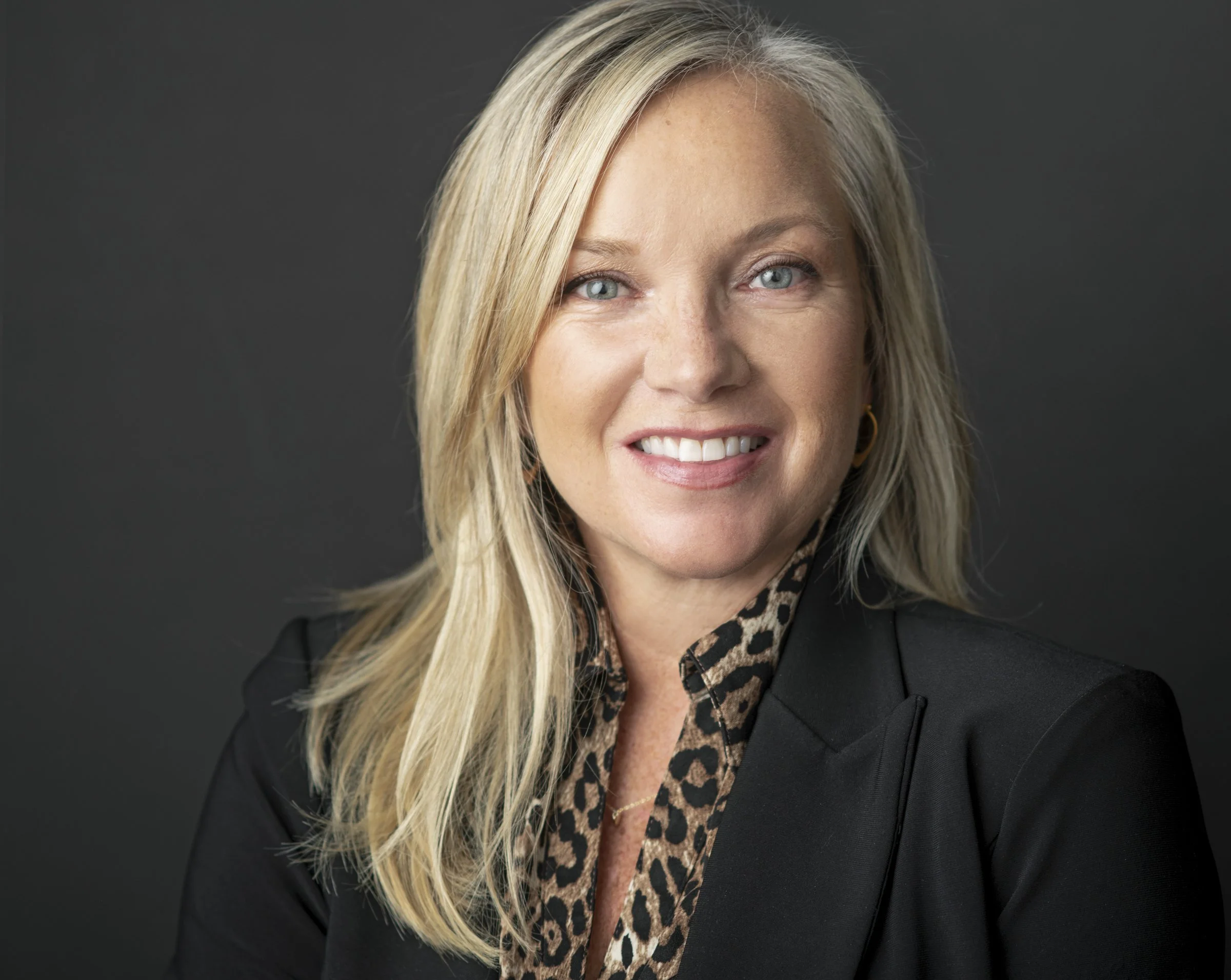 A portrait of a blonde woman with blue eyes smiling, wearing a black blazer, a leopard print shirt, and gold earrings, against a dark gray background.
