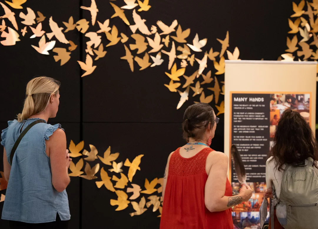 Three women stand in front of an exhibit panel titled 'Many Hands' at a museum or gallery, with yellow paper birds flying on a black background behind them.