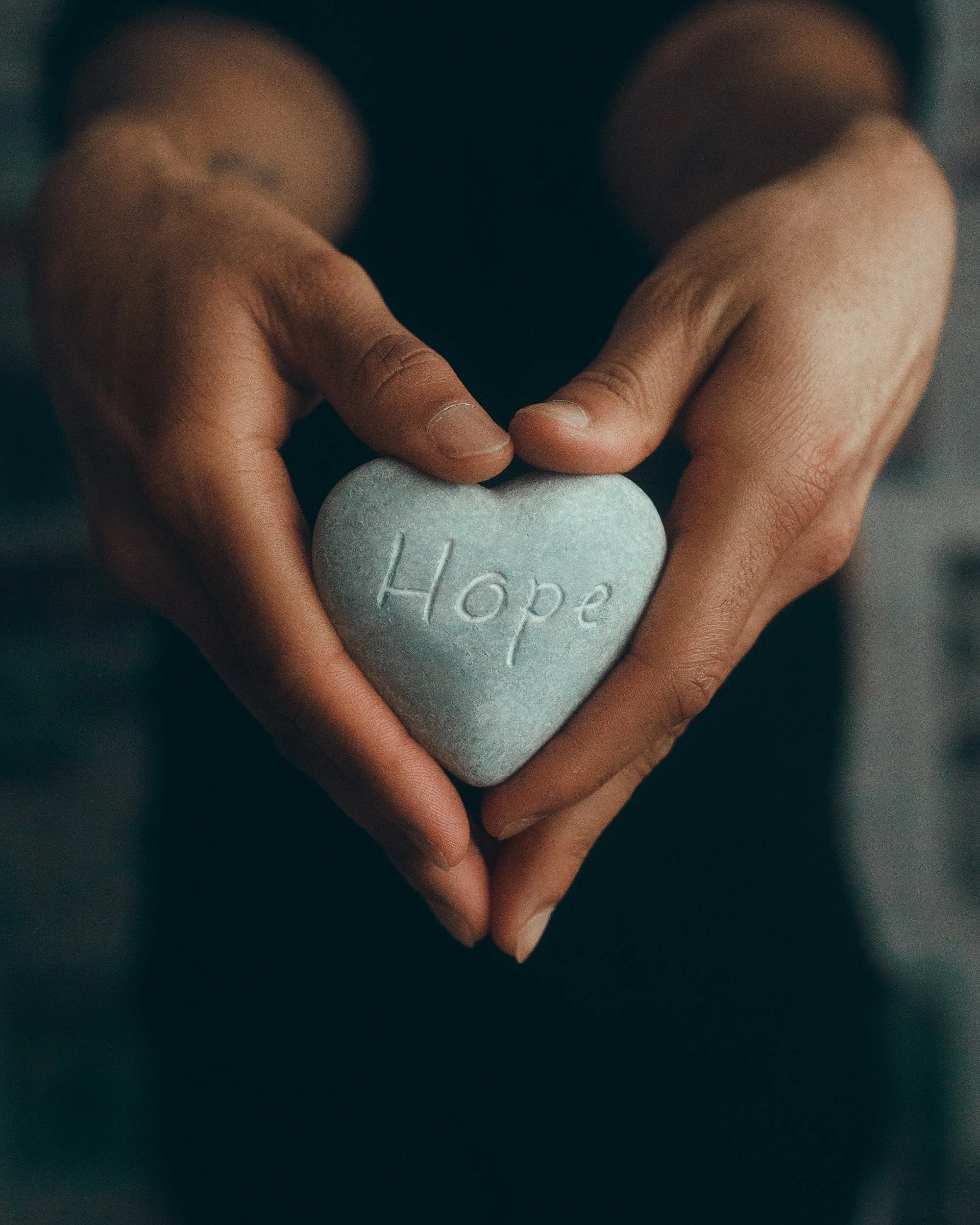 Two hands forming a heart shape around a smooth, gray heart-shaped stone with the word 'Hope' engraved on it.