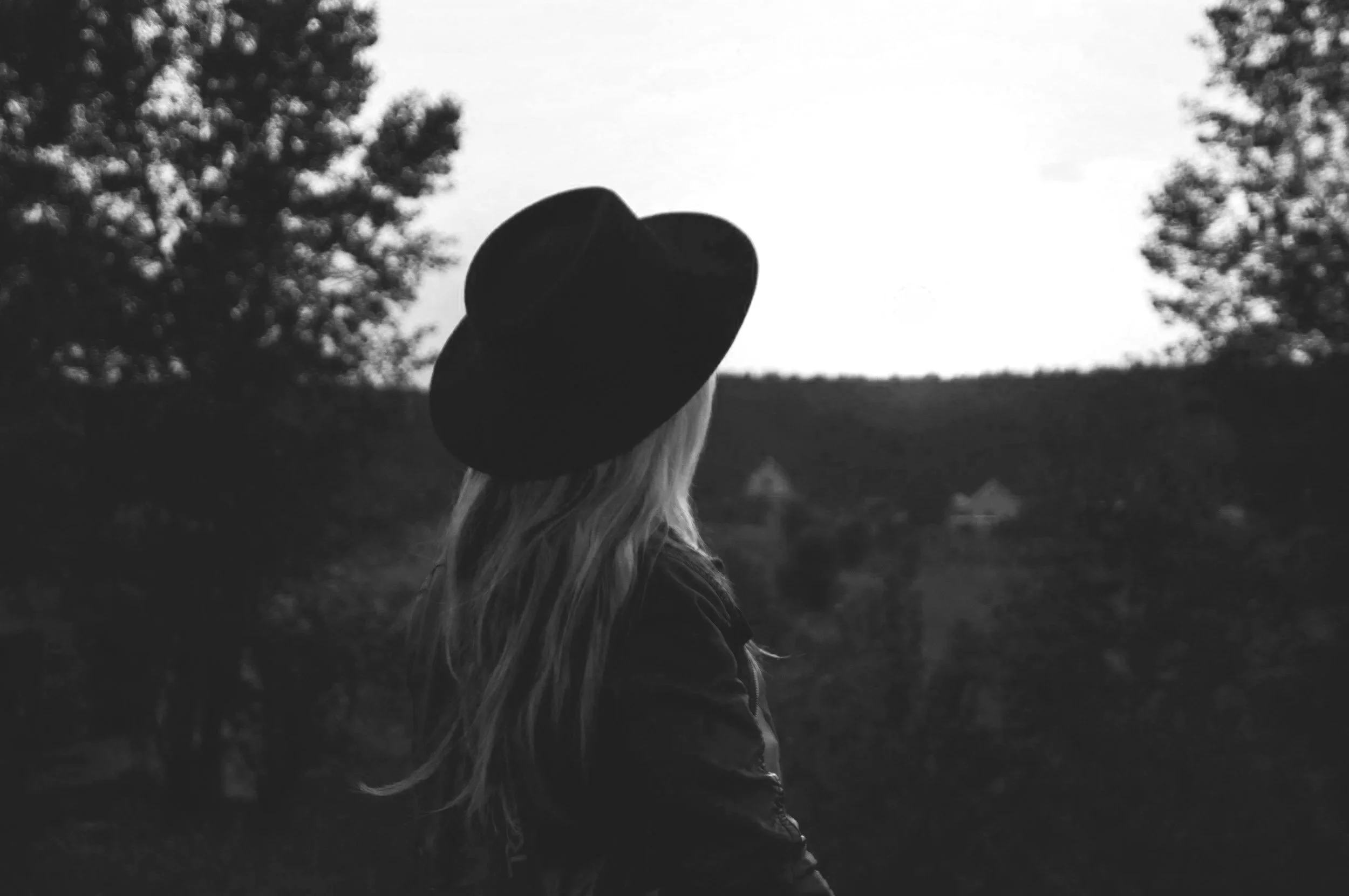A woman with long hair wearing a hat and jacket, looking away in a natural outdoor setting at dusk or dawn, with trees in the background.