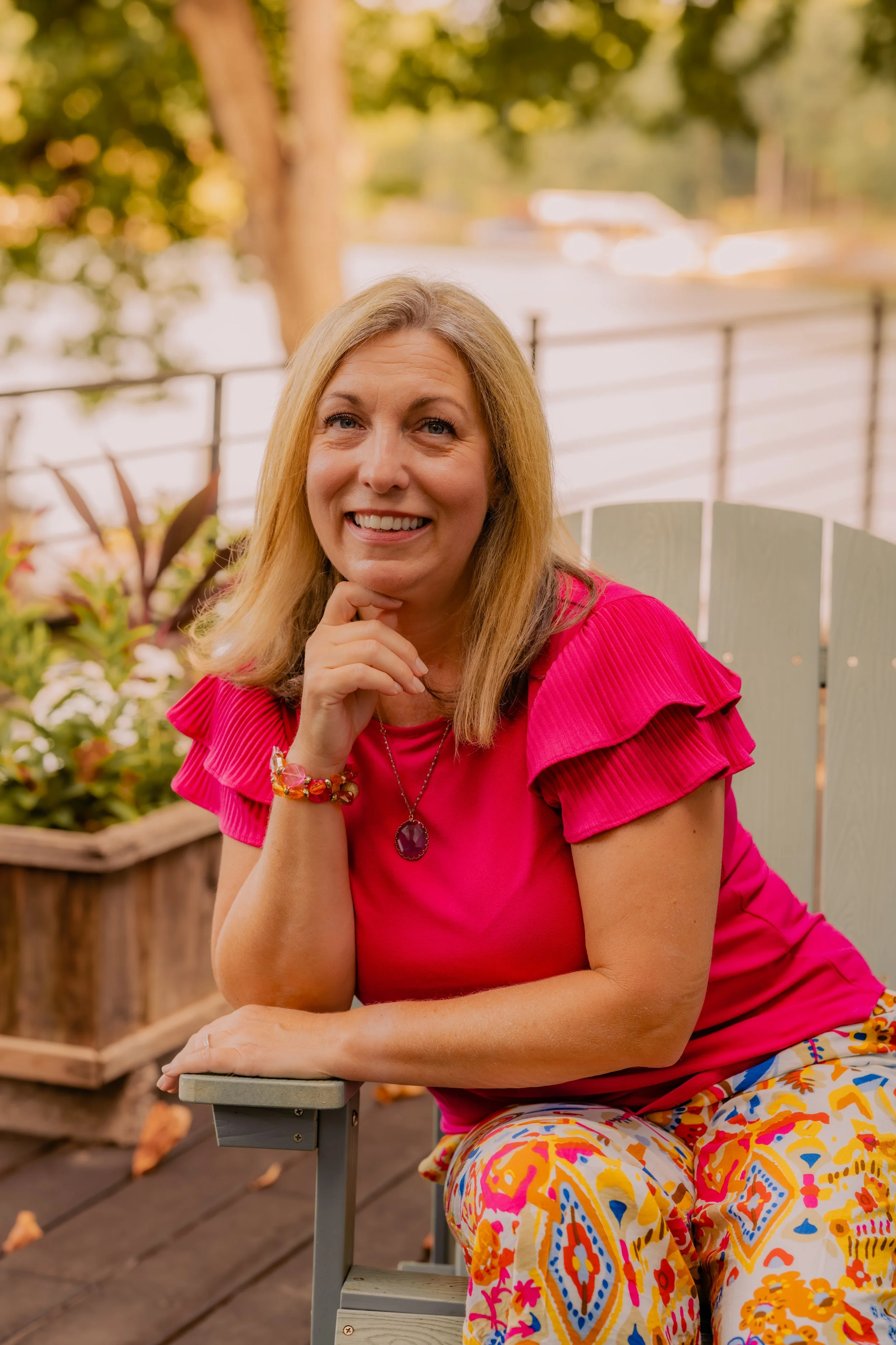 A woman with blonde hair sitting outdoors on a bench, wearing a bright pink top with ruffled sleeves and colorful patterned pants, smiling at the camera.