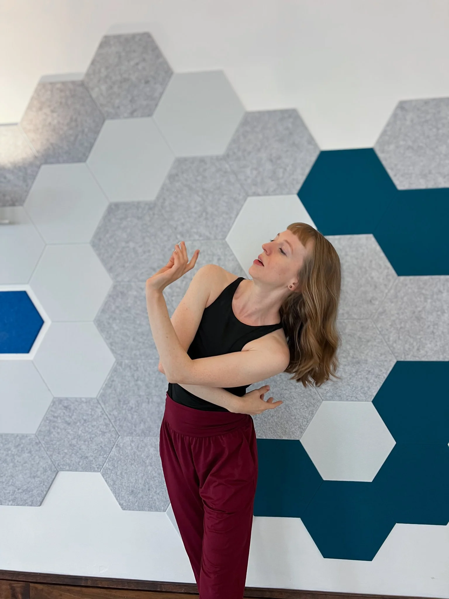 A woman with red hair, wearing a black tank top and maroon pants, standing in front of a wall decorated with hexagonal tiles in shades of gray, white, and blue, striking a dance pose.