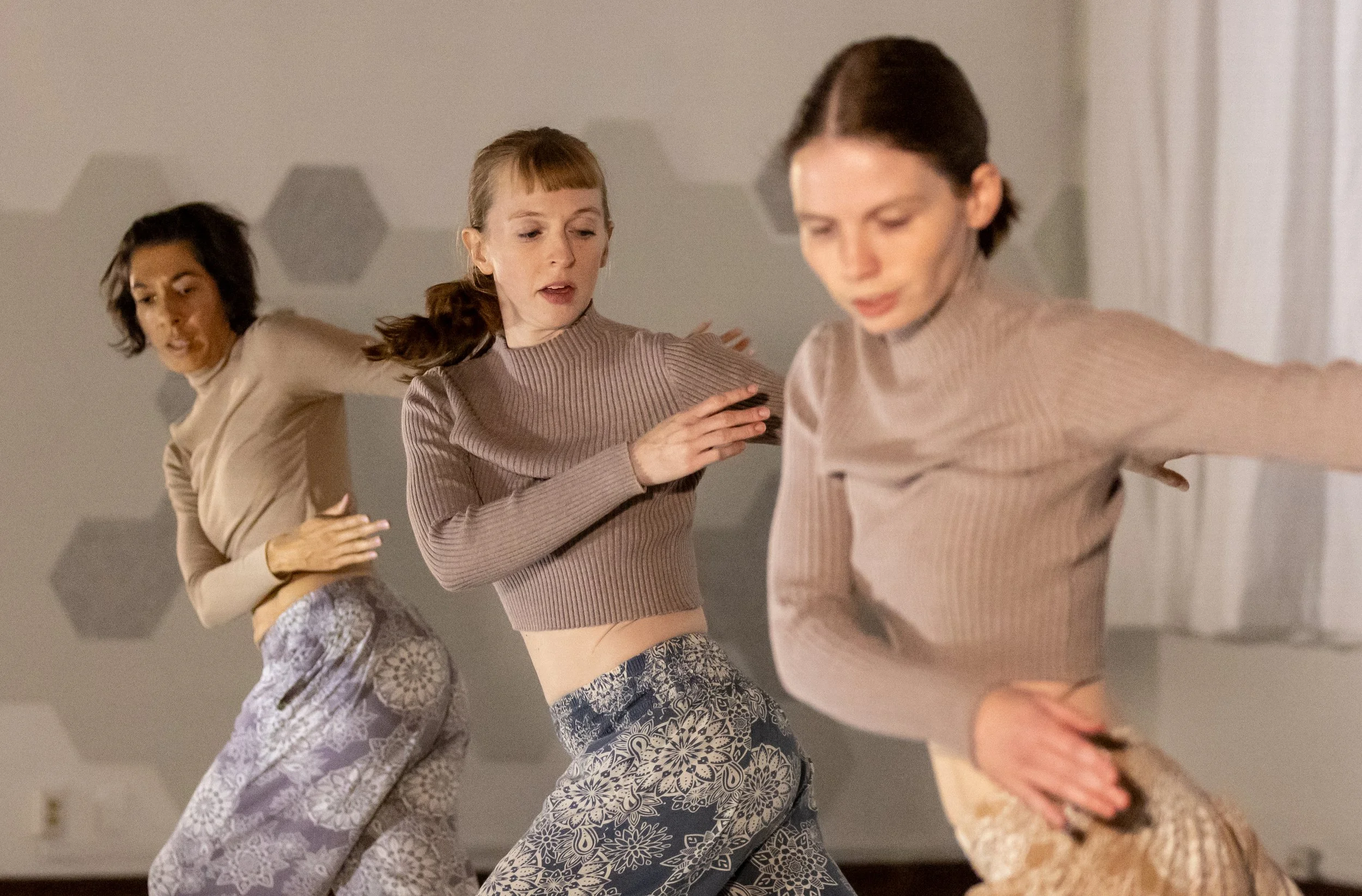 Three women in matching ribbed sweaters and patterned pants perform a contemporary dance in HDC's studio, gaze cast down towards the ground as their arms cross over their bodies.