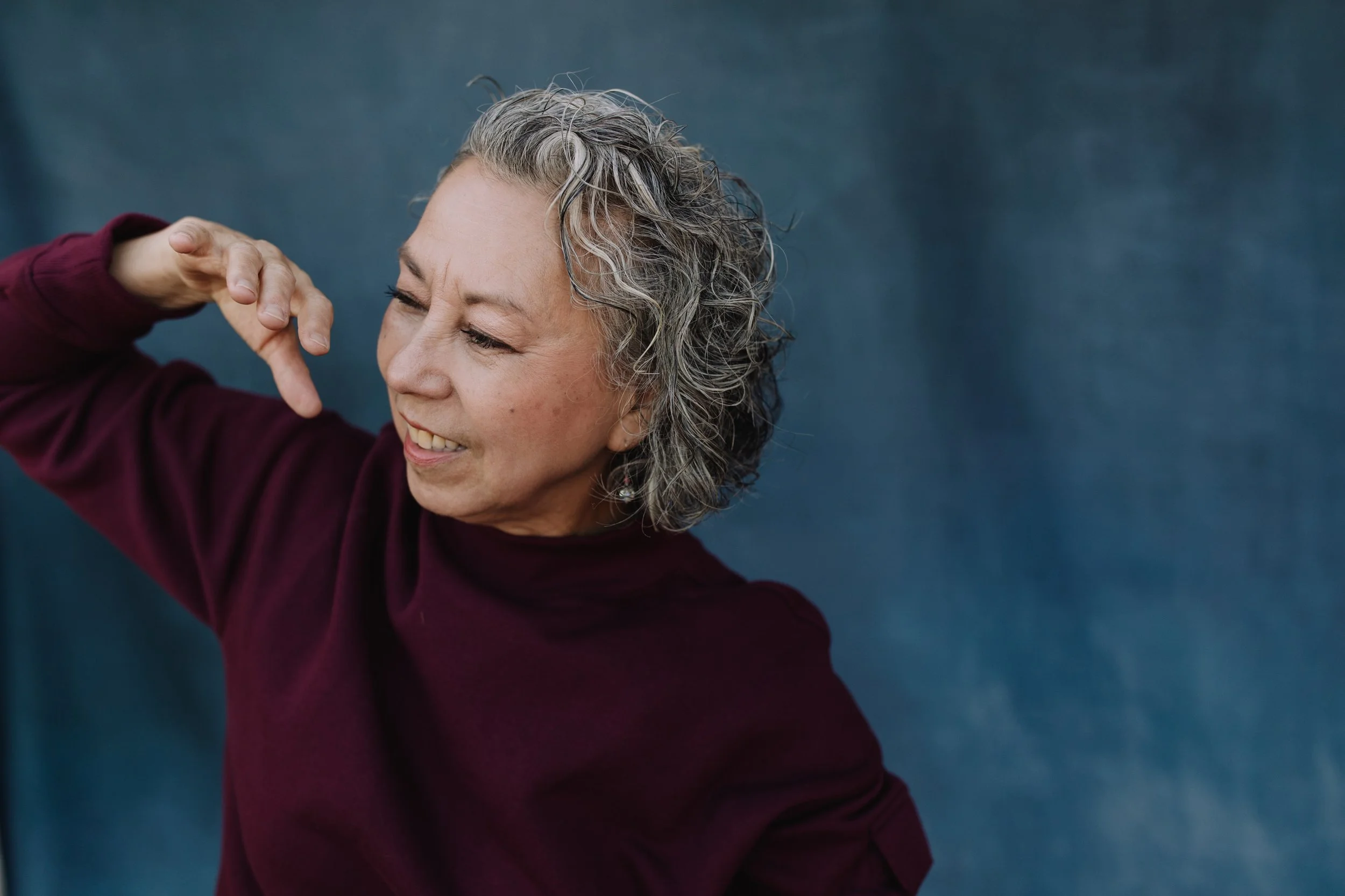 Older woman with gray, curly hair touching her ear, smiling softly, wearing a maroon top, against a dark blue background.