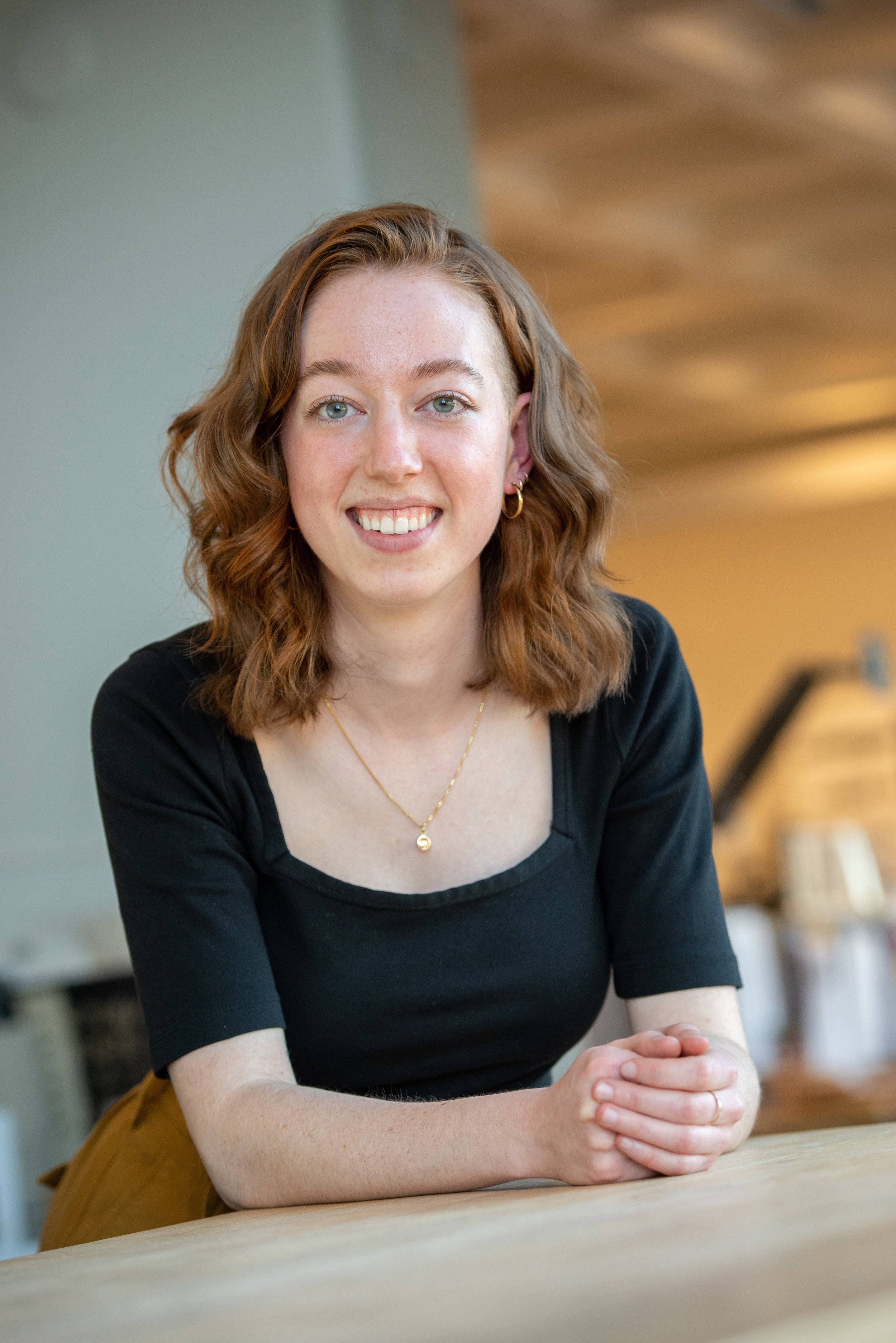 A young woman with shoulder-length curly red hair, wearing a black top, gold jewelry, and smiling, seated at a wooden table in an indoor space with warm lighting.