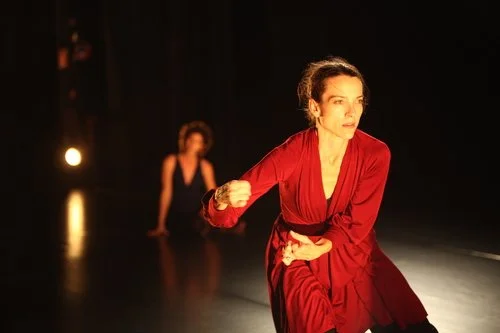 A woman in a red dress performs a modern dance, with another woman in the background on a dark stage with spotlights.