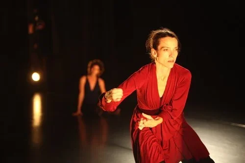 Dancer and choreographer Darla Stanley wears a flowing red dress and performs a dance onstage, set in high contrast against the dark background. She expresses intensity and focus. Another performer sits out of focus behind her.