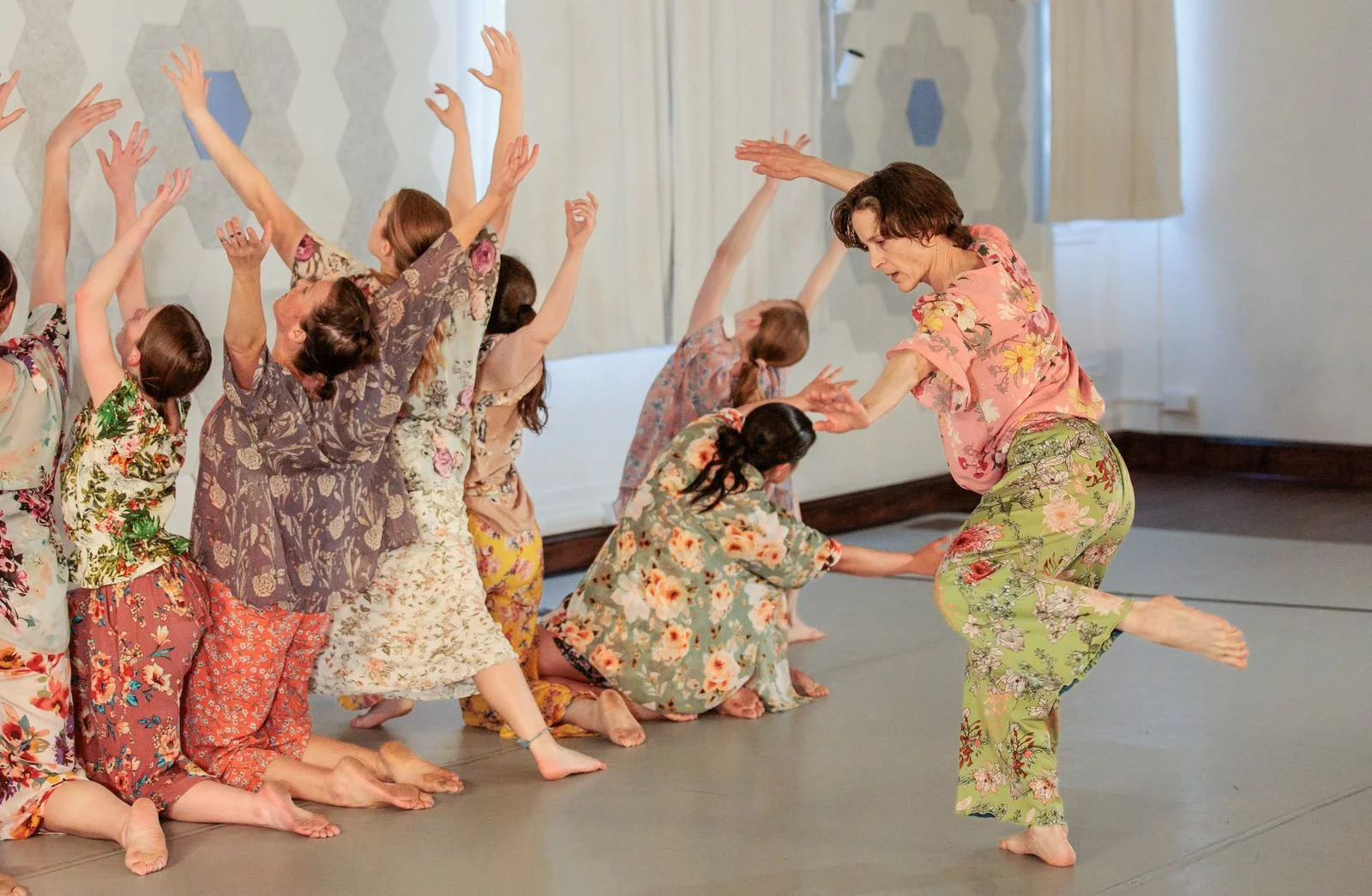 Dancers in bright, floral clothing perform an intense contemporary dance piece, with one dancer moving across the stage while the others kneel behind her, reaching up towards the ceiling.