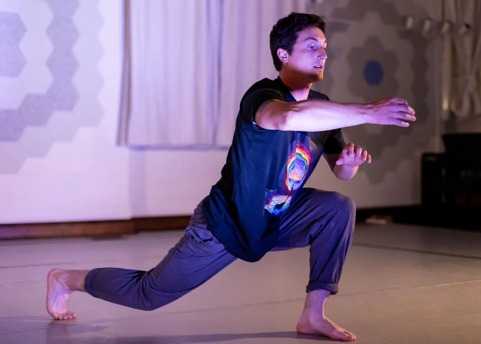 A man in a black T-shirt with a colorful rainbow design, brown pants, and barefoot is performing a martial arts stance indoors, with one knee bent forward, the other leg extended back, and arms outstretched.