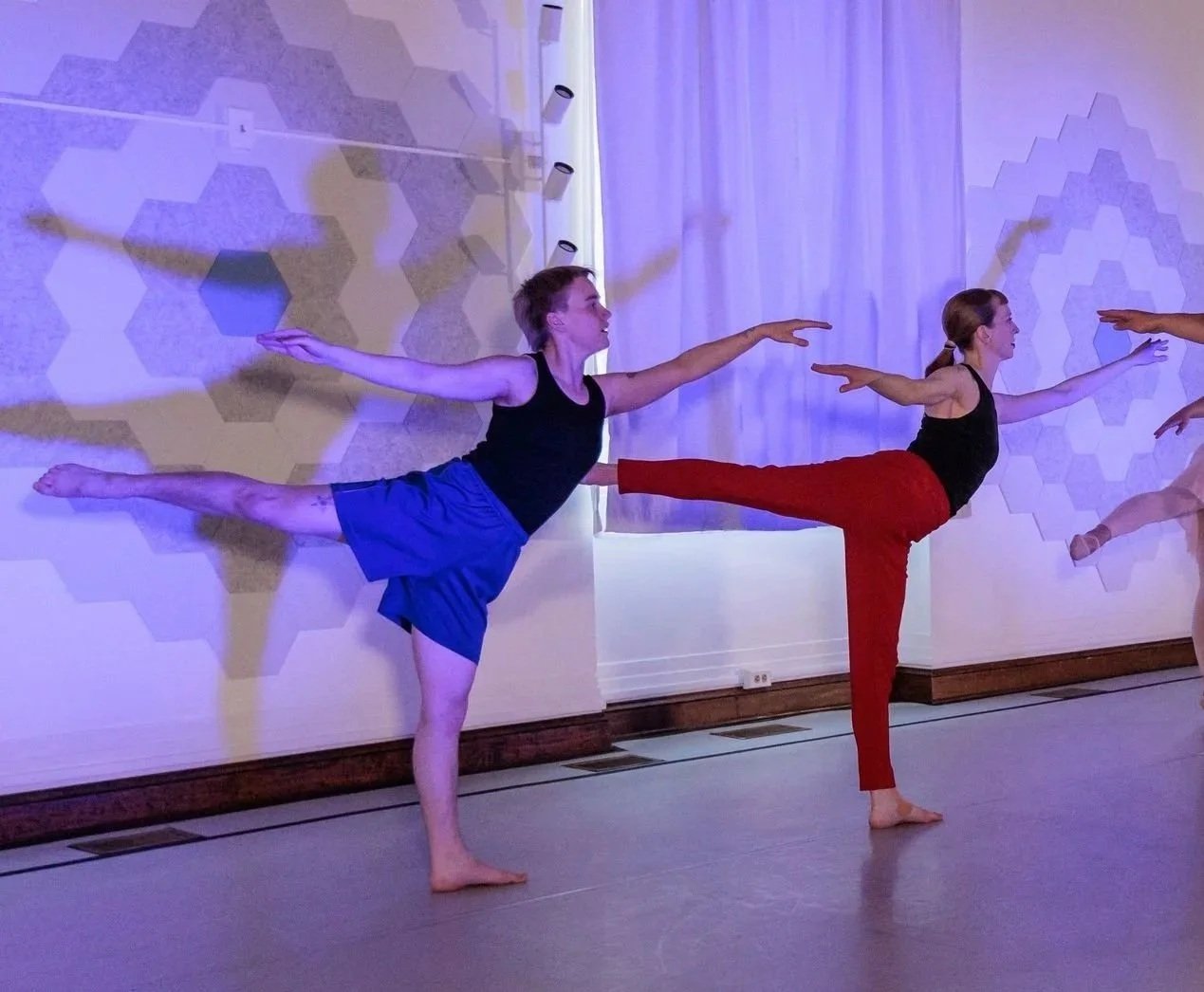 Two dancers perform an arabesque position with their back legs lifted and their front arms reaching forward. Stage lighting creates distinct dark shadows on the wall behind them.