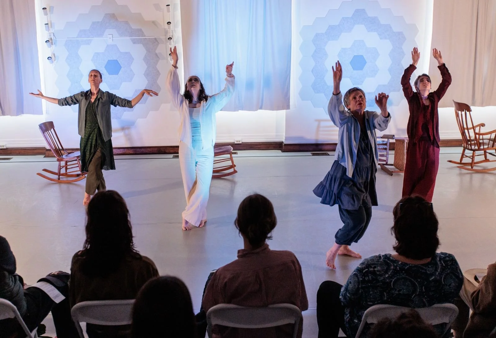 Four dancers on stage with their arms raised, in front of an audience, with rocking chairs on stage and geometric patterned backdrop.