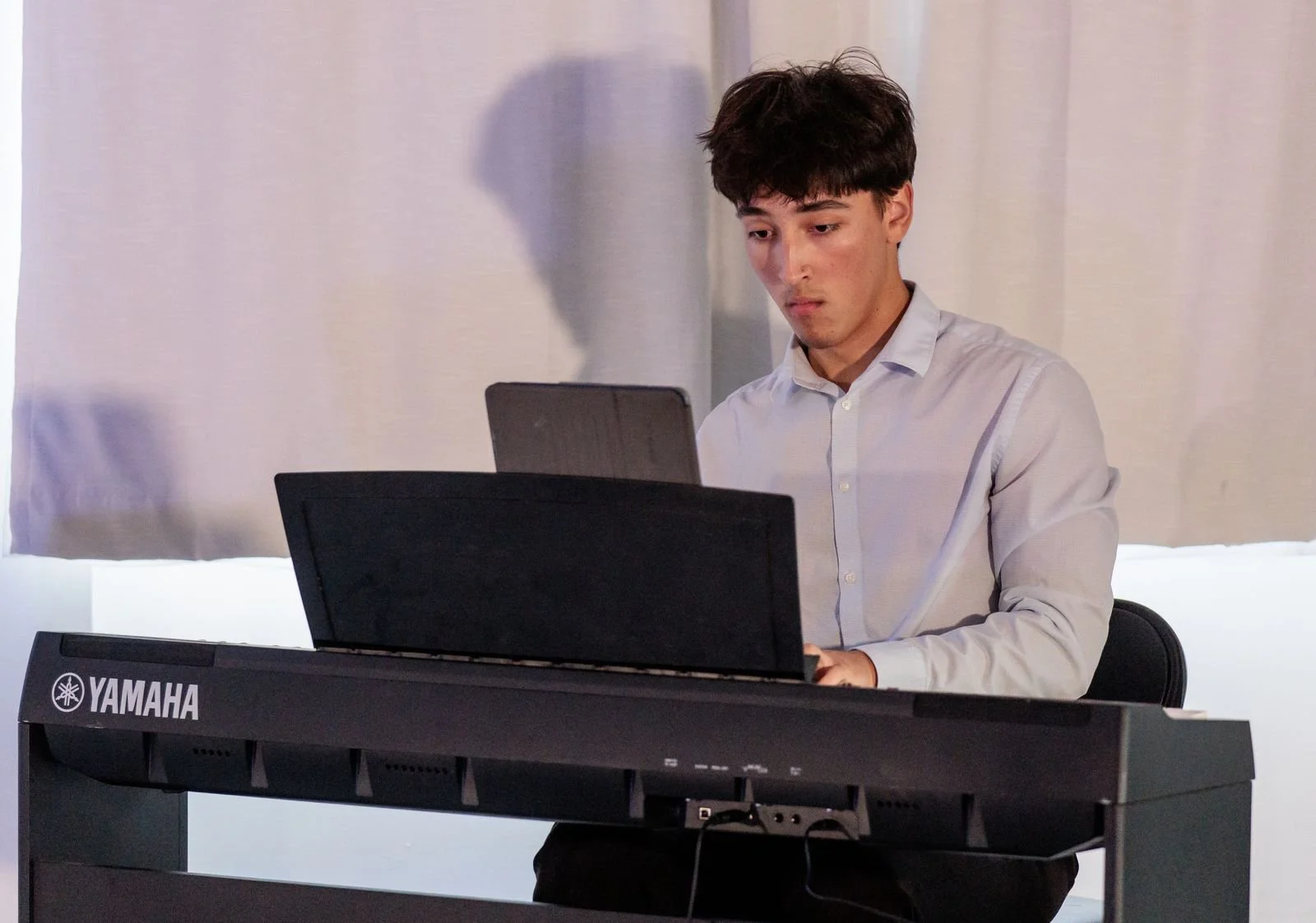 A young man with dark hair wearing a white button-up shirt plays a Yamaha keyboard in a room with plain white curtains.