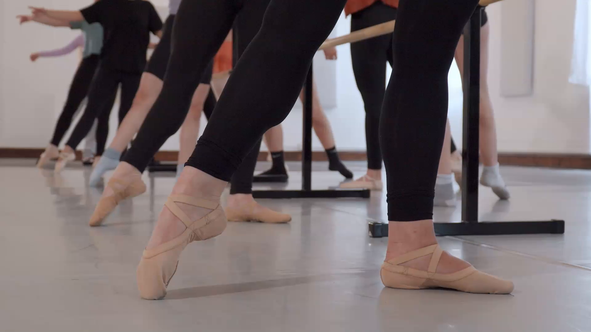 Ballet dancers at the barre in a busy ballet class with their feet pointed, shown from the knee down. The dancers wear black tights and ballet slippers.