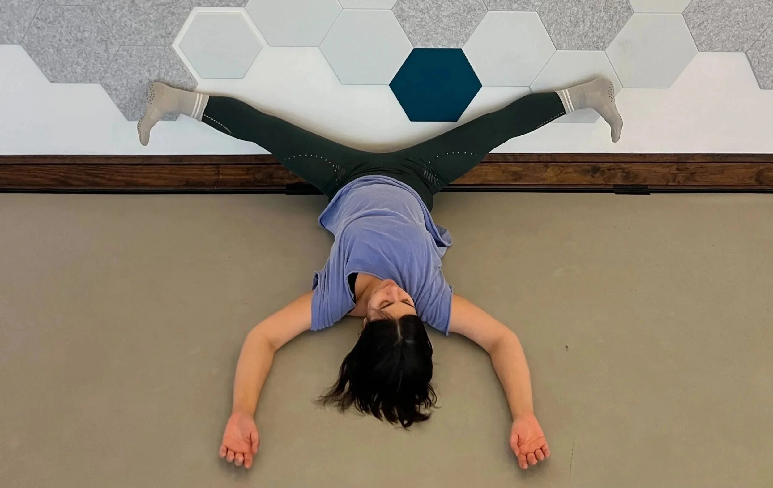 A dancer lies on the floor, stretching her legs wide against the geometrically patterned wall of the White Box Studio.