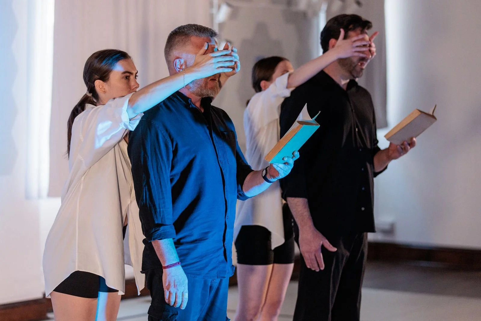 Two men in black clothing read poetry aloud from old books, each with their eyes covered by the hands of a dancer standing behind them. The dancers wear white shirts.