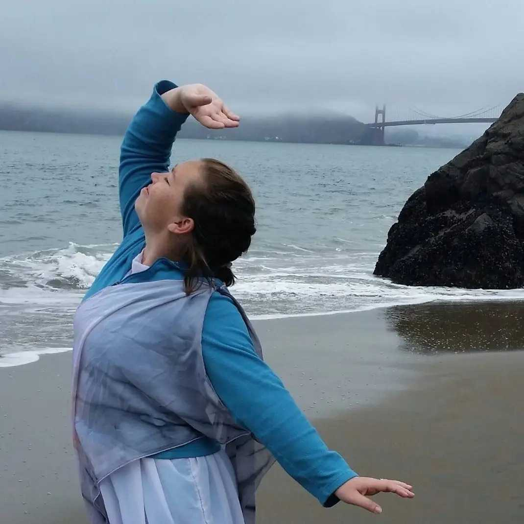 A woman practicing yoga on a beach with waves, a large rock, foggy weather, and the Golden Gate Bridge in the background.