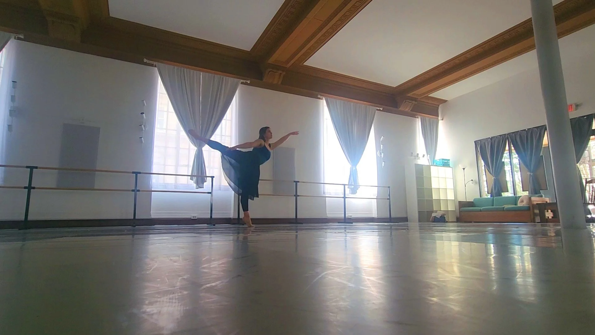 A woman in a dance studio holding an arabesque pose, standing on one leg with her other leg extended behind her and arms outstretched, wearing a teal top and long black skirt.